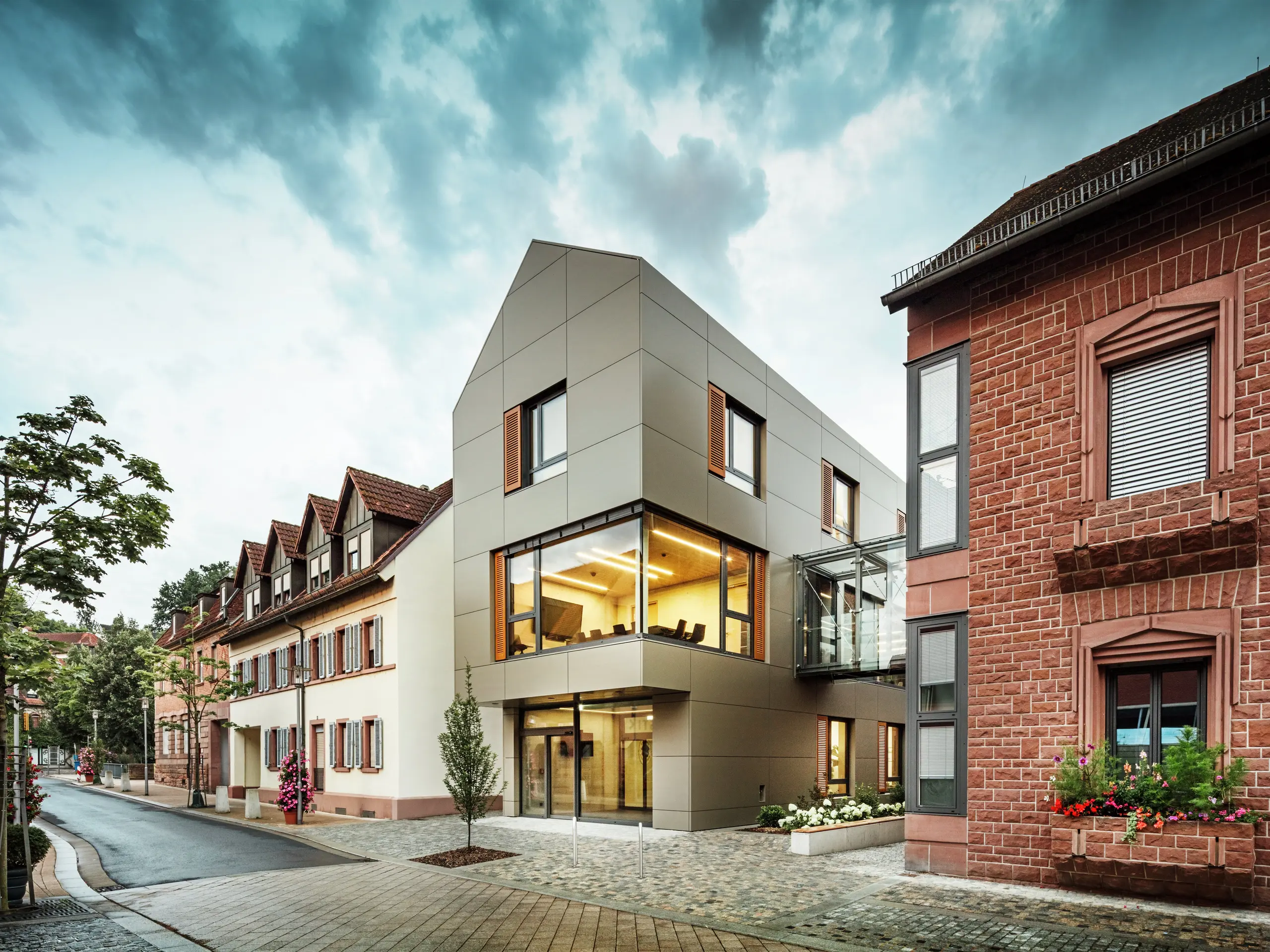 Das Bild zeigt das moderne Rathaus in Alzenau, dessen Fassade mit PREFA-Aluminiumverbundplatten in einem warmen Bronzeton verkleidet ist. Große Glasflächen unterbrechen die Fassade und geben den Blick auf ein lichtdurchflutetes Interieur frei. Ein gläserner Verbindungsgang auf der rechten Bildseite verbindet das Gebäude mit einem nahegelegenen traditionellen Backsteingebäude und schafft so einen interessanten Kontrast zwischen moderner und historischer Architektur.