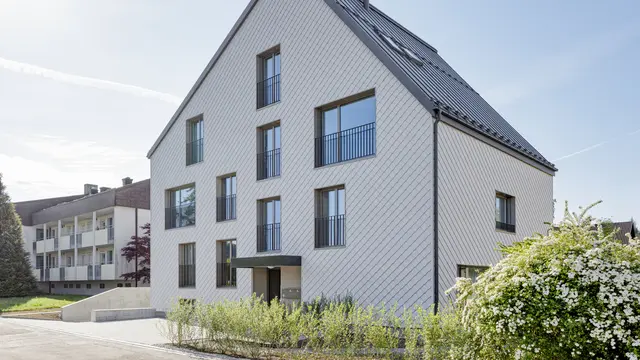Modernes Mehrfamilienhaus in der Schweiz mit einem langlebigen PREFA Dach aus PREFALZ in P.10 Dunkelgrau und einer Fassade, verkleidet mit PREFA Wandrauten 20x20 in P.10 Prefaweiß. Das Objekt bietet eine zeitgemäße Architektur, die durch die Kombination von traditionellem Satteldach und minimalistischen Fassadenelementen besticht. Die klare Linienführung und das strukturierte Erscheinungsbild der Wandrauten sind harmonisch in eine gepflegte Umgebung mit üppigem Blütenstrauch integriert und zeigen die Vielseitigkeit von Aluminium als Baumaterial.