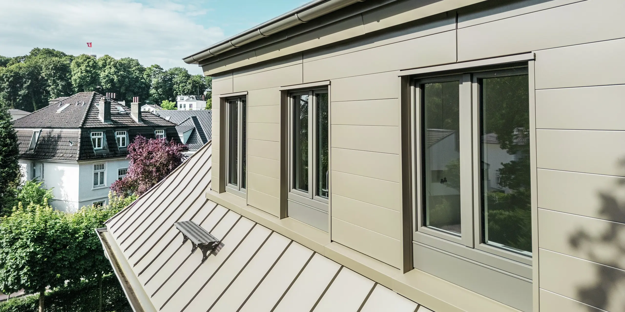 Detailansicht der Villa Blankenese in Hamburg mit edlem PREFALZ Dach und PREFA Sidings in Bronze. Das Bild zeigt die klare Linienführung und die hochwertige Aluminiumverarbeitung, die für Langlebigkeit und Witterungsbeständigkeit sorgt. Die bronzene Oberfläche verleiht dem Gebäude eine elegante, moderne Optik und setzt markante Akzente im Zusammenspiel mit den großen Fenstern. Die PREFA Produkte bieten nicht nur stilvolles Design, sondern auch wartungsfreie Qualität – perfekt für anspruchsvolle Architektur in jeder Umgebung.
