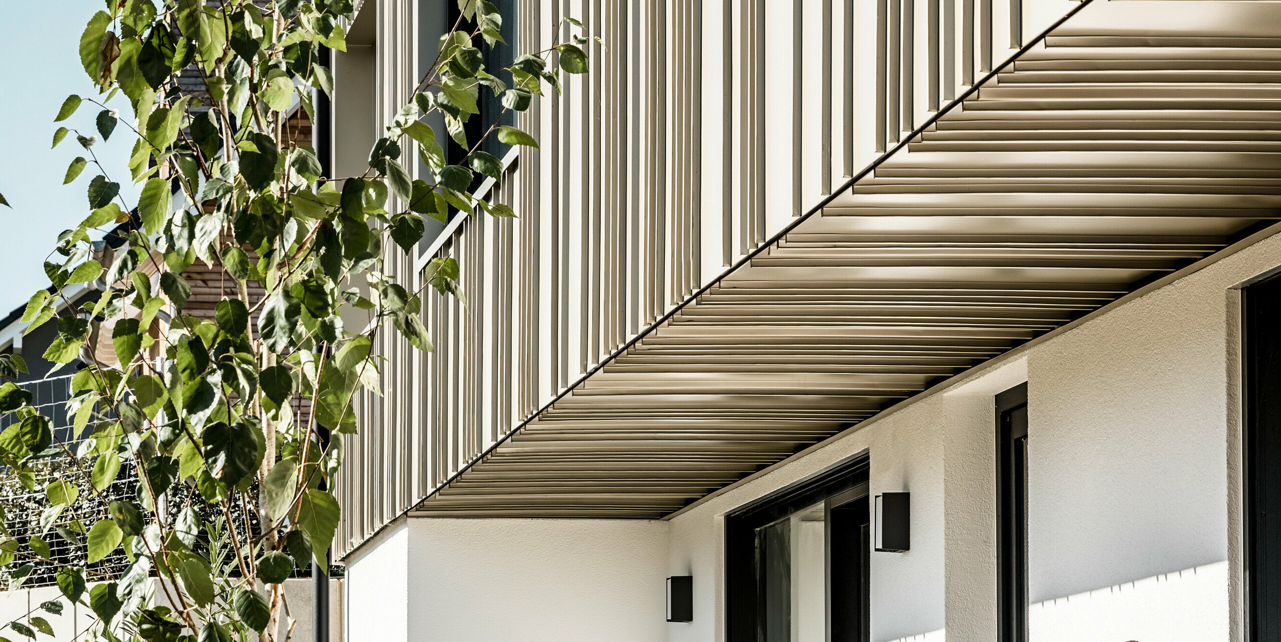 Seitenansicht eines zeitgenössischen Hauses mit einem PREFA Fassadensystem in Lichtbronze, das eine präzise Verarbeitung zeigt. Die Aluminiumfassade aus FALZONAL bildet einen attraktiven Kontrast zum schlichten Weiß des Erdgeschosses. Die dunklen Fensterrahmen und Außenlichter betonen die klare Linienführung der Architektur. Im Vordergrund verleihen grüne Bäume dem Bild eine natürliche Tiefe und unterstreichen die umweltfreundliche Bauweise mit langlebigen und recyclebaren PREFA Aluminiumprodukten.