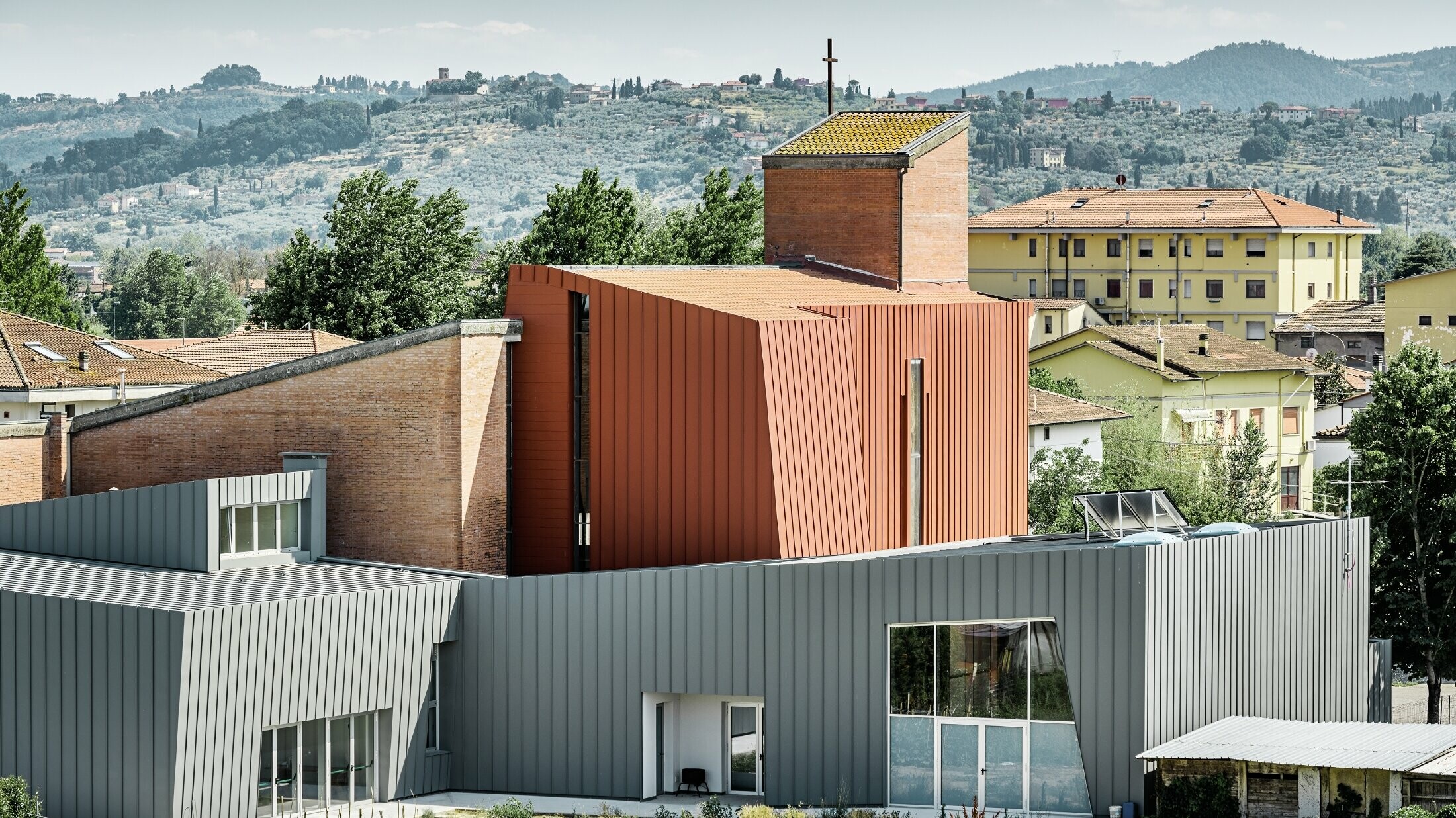 Modernes Kirchengebäude mit markanter Architektur, bestehend aus klaren geometrischen Formen. Der Bau kombiniert hellgraue Aluminiumfassaden mit einem zentralen, ziegelroten Turm aus Metallpaneelen und Ziegelmauerwerk, auf dessen Spitze ein Kreuz steht. Im Hintergrund sind Wohnhäuser und eine hügelige, grüne Landschaft mit Bäumen und Olivenhainen zu sehen.