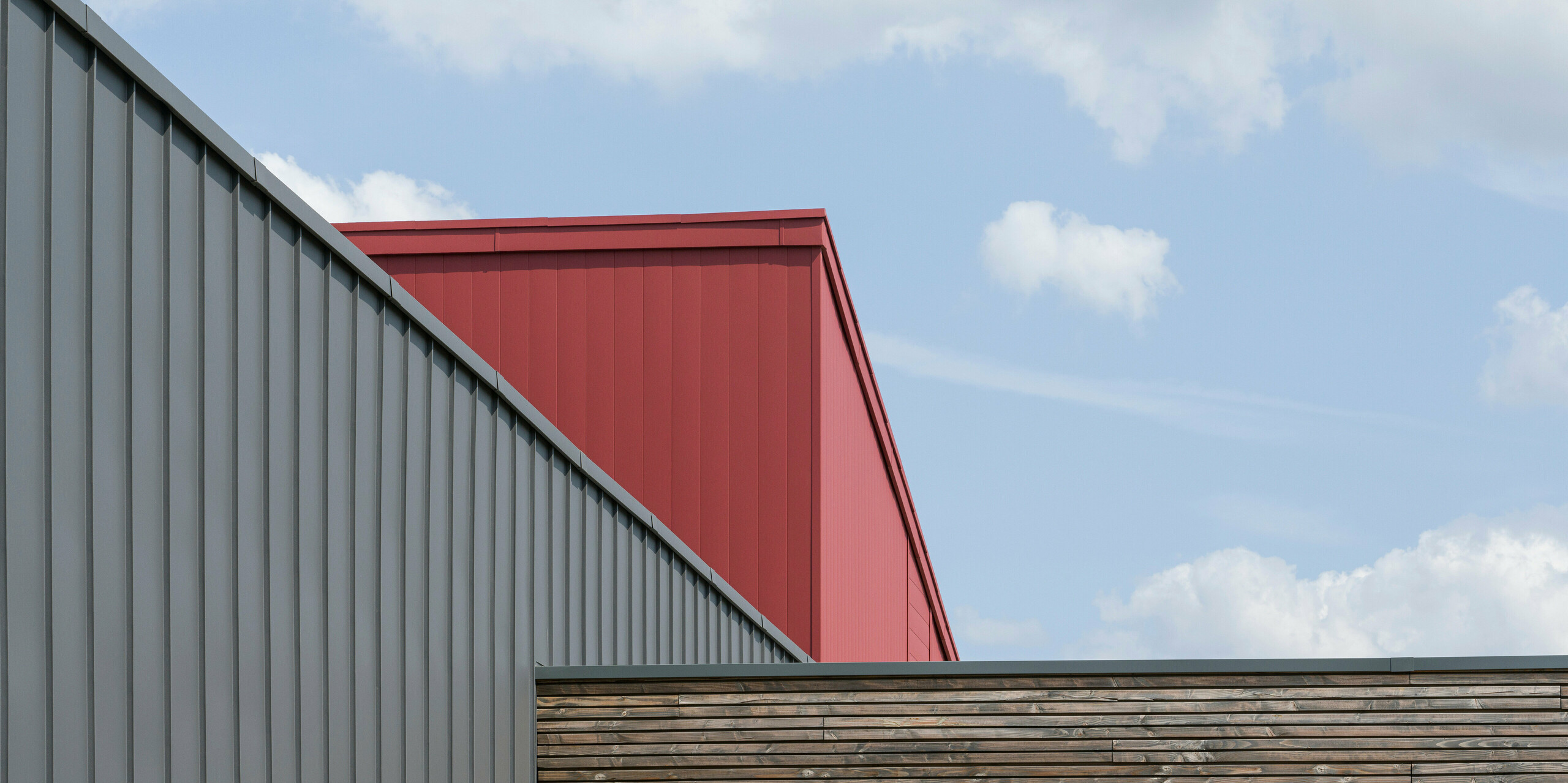 Nahaufnahme der Dach- und Fassadengestaltung eines modernen Gebäudes. Im Vordergrund ist eine Wand mit horizontaler Holzverkleidung zu sehen, die eine natürliche Textur aufweist. Dahinter erhebt sich eine dunkelgraue PREFALZ-Fassade mit vertikalen Scharen, die eine industrielle Eleganz ausstrahlt. Über dieser Fassade zeigt sich ein Ausschnitt eines leuchtend roten Dach- und Fassadensystems aus Aluminium, das einen auffallenden Farbkontrast bietet und die moderne Architektur unterstreicht.