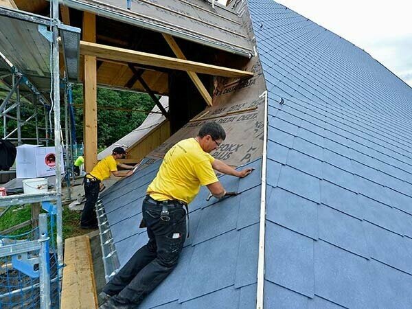 zwei Handwerker bei der Sanierung des Daches am Fusenhof mit gelben T-Shirts, sie Decken das Dach des alten Bauerhofes mit der PREFA Aluminium Dachraute in steingrau ein, darunter liegt eine graue Folie