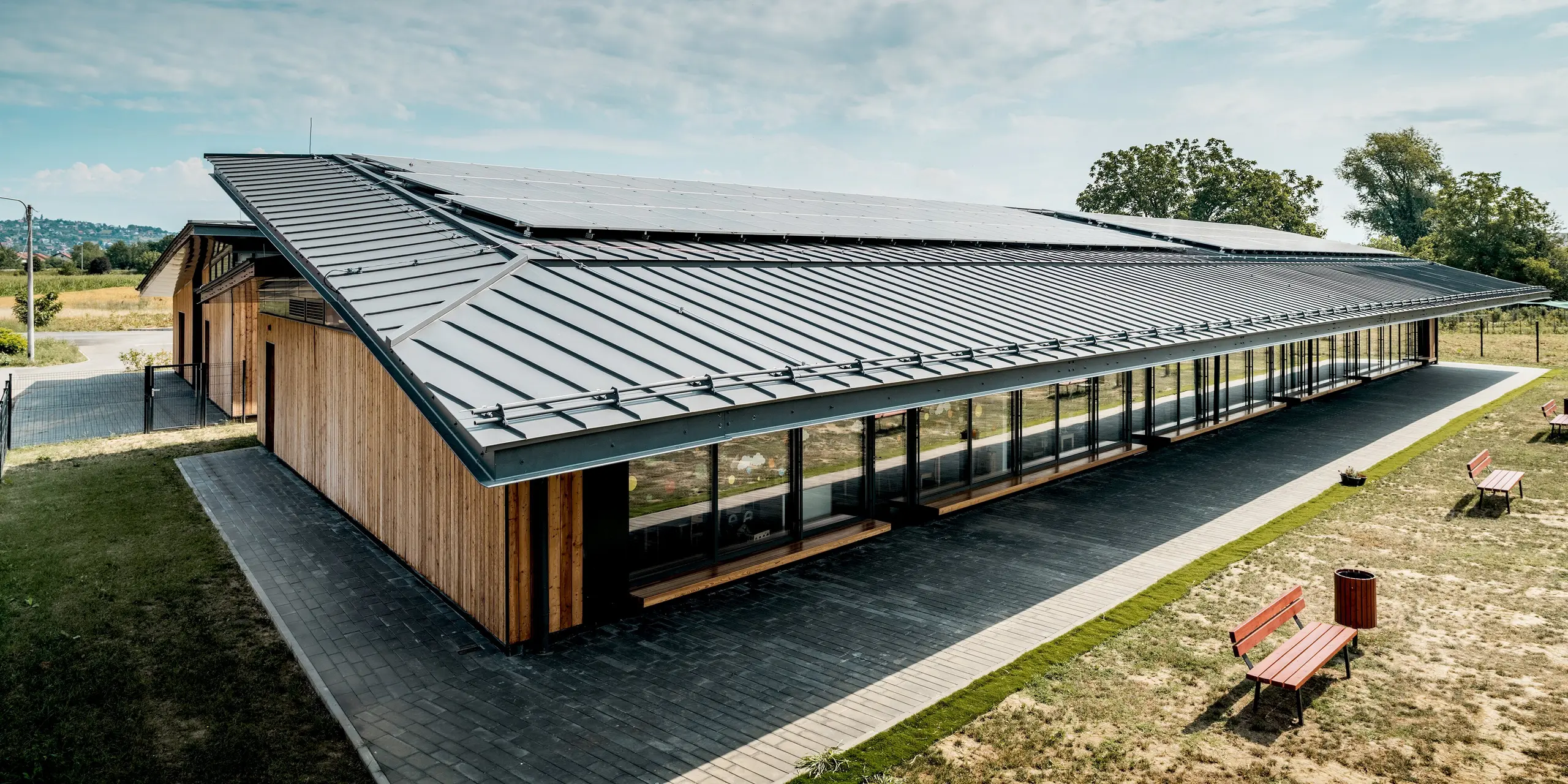 Weitläufige Ansicht des Kindergartens Svarozic mit markantem, weit auskragendem PREFALZ Aluminiumdach in P.10 Anthrazit. Die elegante Dachfläche überspannt die großzügigen Glasfronten und die natürliche Holzfassade, während sie mit klaren Linien die moderne Architektur betont. PREFA Aluminium sorgt für langlebigen Schutz, hohe Widerstandsfähigkeit und eine zeitlose Optik.