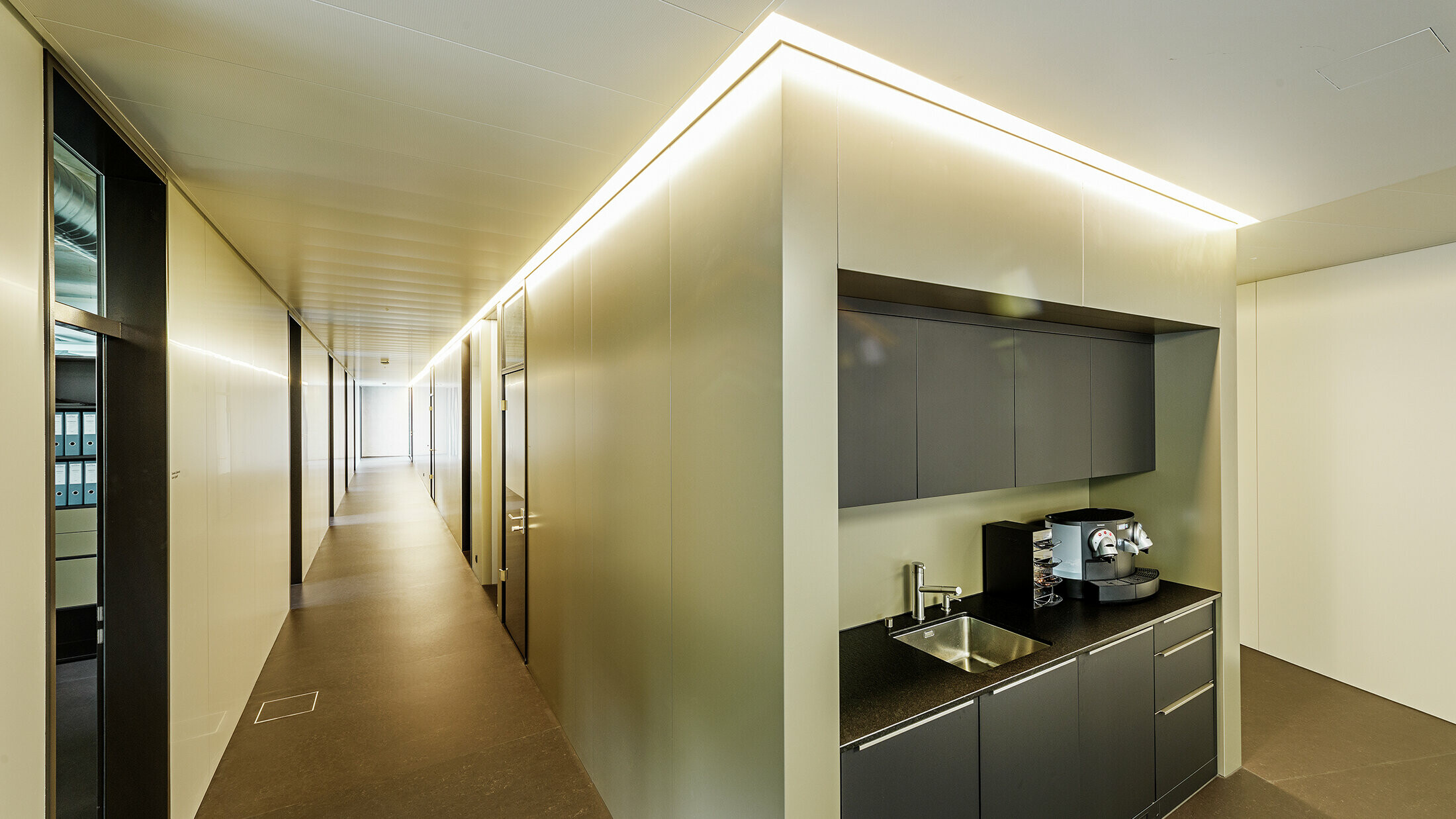 In den modernen Büroräumlichkeiten der Firma Guggisberg Dachtechnik in Wabern sorgt die integrierte Teeküche für Funktionalität und Design. Die Wandverkleidungen nehmen die edle Bronzeoptik der PREFA Aluminiumfassade auf und fügen sich nahtlos in das architektonische Konzept ein. Die Innenraumgestaltung mit indirekter Beleuchtung schafft ein angenehmes Raumgefühl.