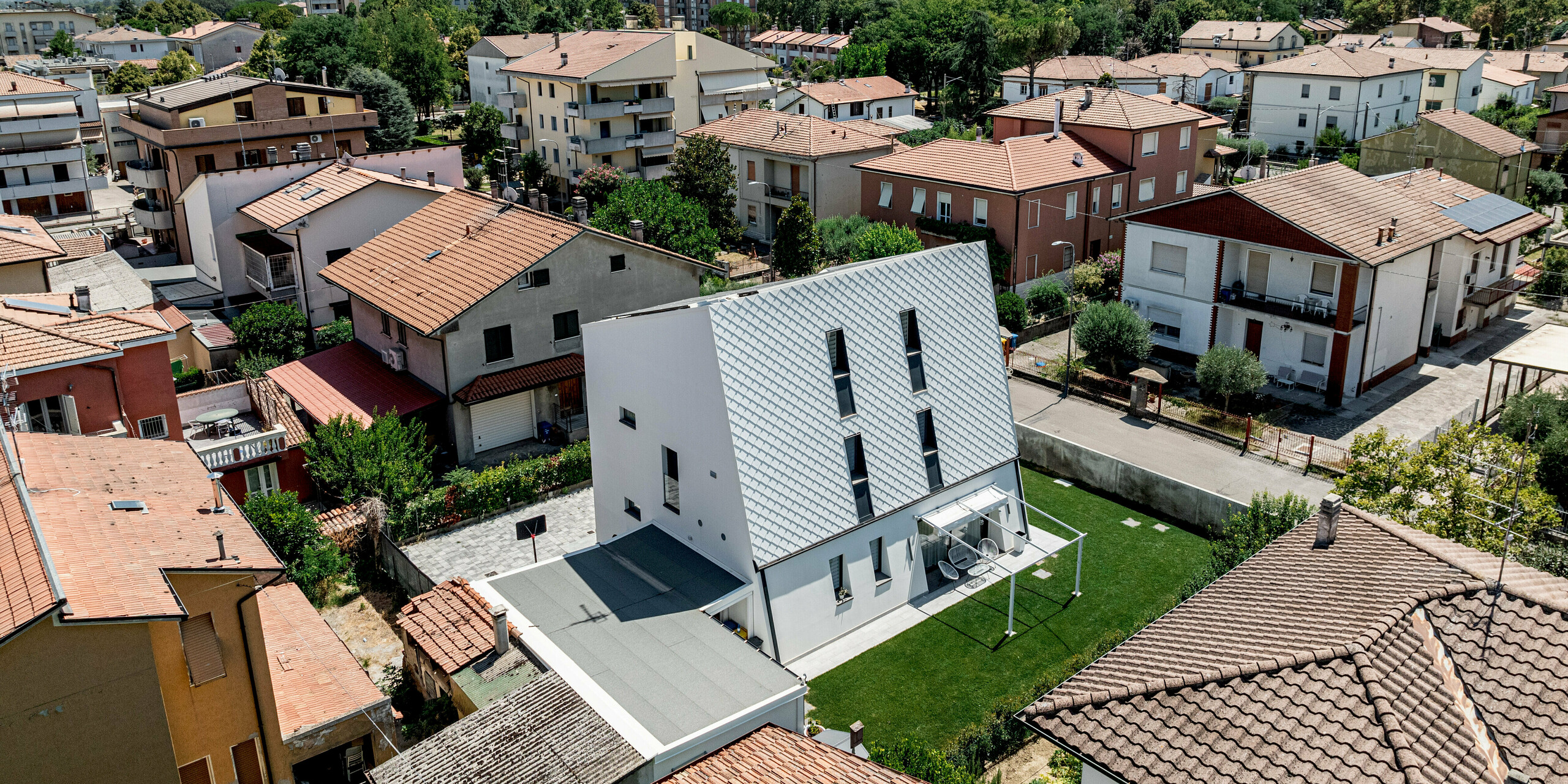 Luftaufnahme eines modernen Einfamilienhauses inmitten eines dicht bebauten Wohngebiets in Forlì, Italien. Die strahlend weiße, geneigte Hauptfassade aus PREFA Wandraute 29 × 29 in P.10 Reinweiß hebt sich deutlich von den umliegenden Ziegeldächern ab. Das präzise Rautenmuster aus Aluminium bildet einen klaren architektonischen Eyecatcher im Stadtbild und zeigt seine Vorteile: langlebig, witterungsbeständig und nahezu wartungsfrei.