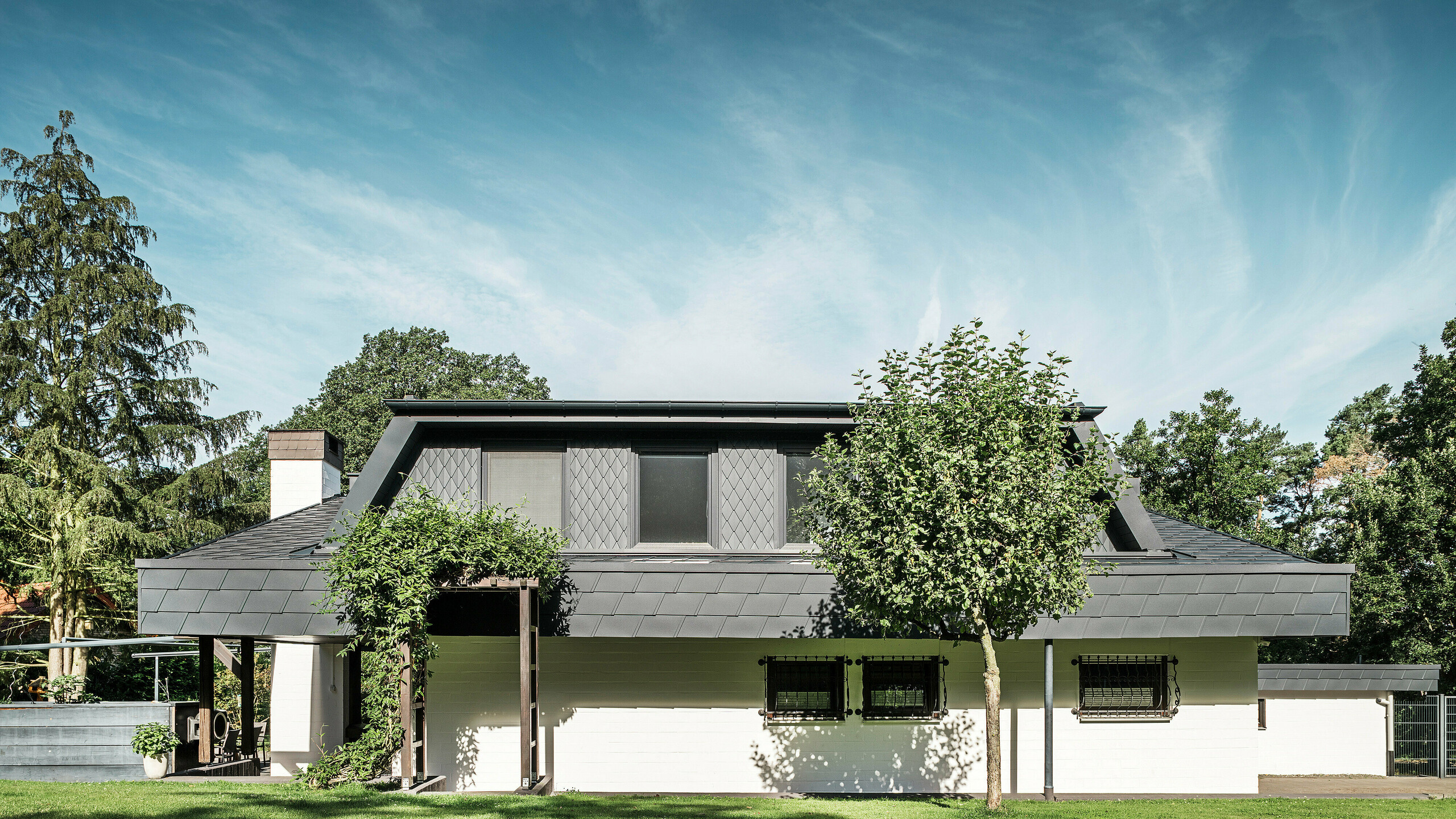 Frontale Ansicht auf die Gartenseite eines Einfamilienhauses in Mechernich mit einer großen Trapezgaube, eingedeckt mit kleinen PREFA Dachrauten P.10 Anthrazit. Das Dach ist mit PREFA Dachschindeln DS.19 im gleichen Farbton eingedeckt. Die Fassade des unteren Geschosses ist weiß gestrichen und eine begrünte Pergola rundet das Gesamtbild im Gartenbereich ab. Bäume und Sträucher rahmen den Blick und sorgen für eine harmonische Einbindung in die Natur.