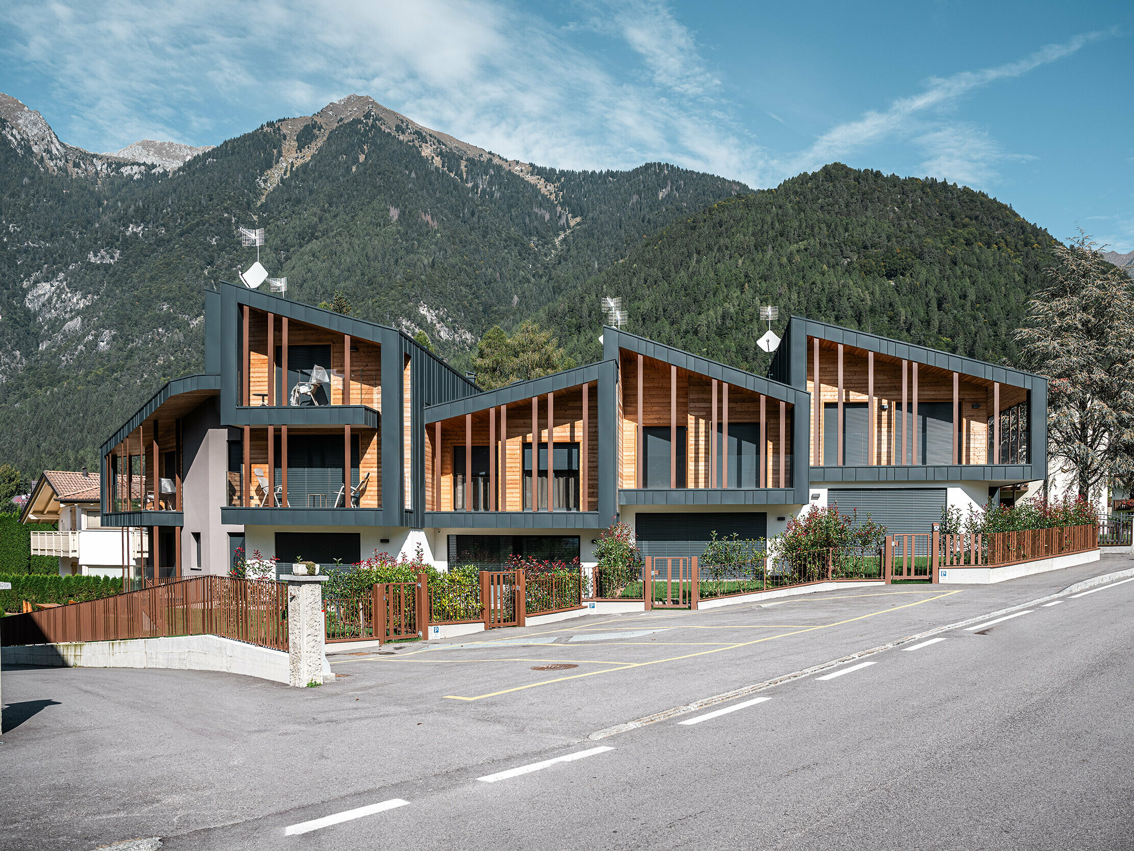 Moderne Ferien-Chalets mit anthrazitfarbenen Prefalz-Metalldächern und Holzelementen in einem alpinen Tal, an einer Straße gelegen, mit umzäunten Vorgärten und einem beeindruckenden Bergpanorama im Hintergrund.