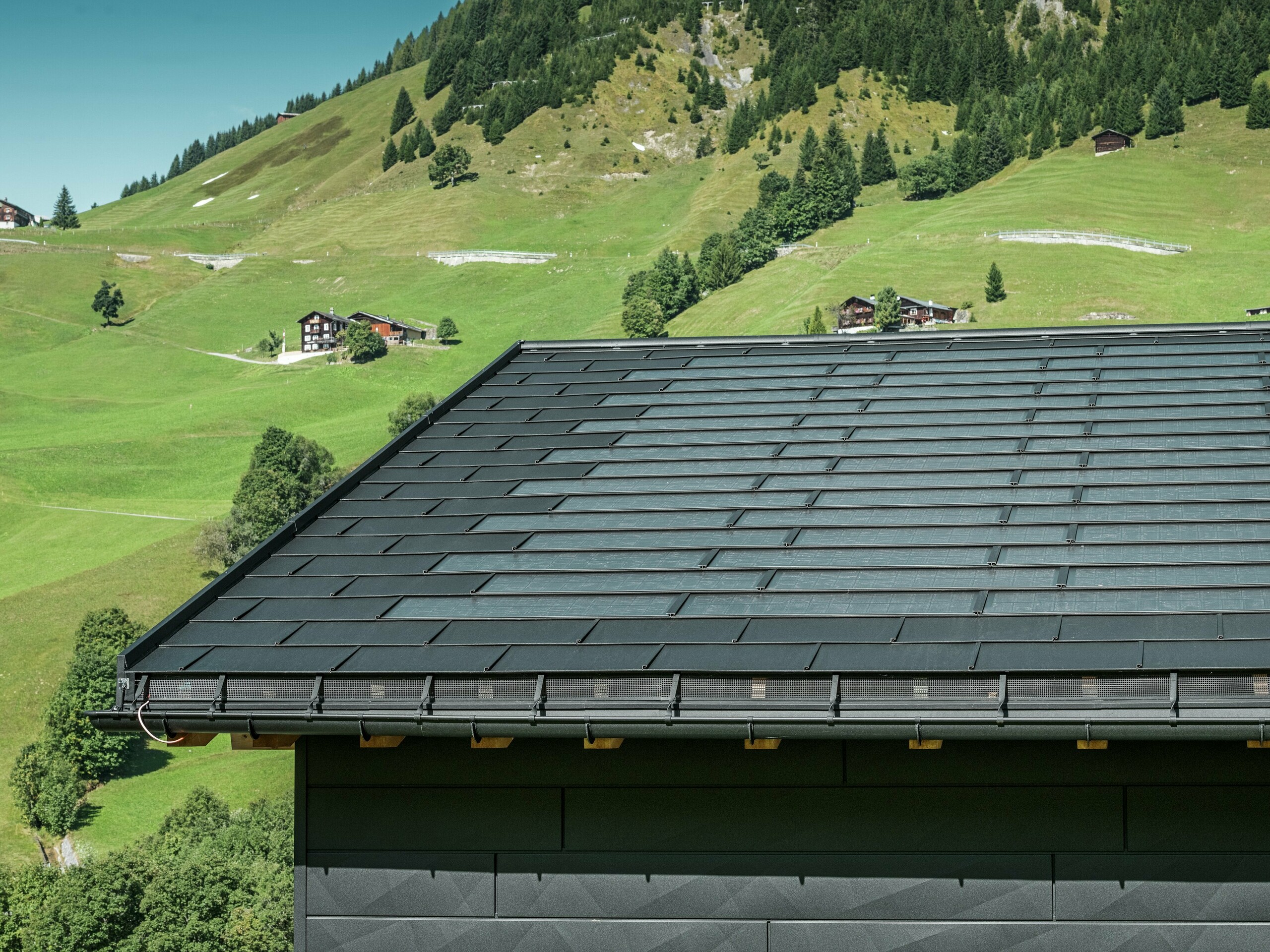 Nahaufnahme der Traufe eines modernen Einfamilienhauses in der alpinen Umgebung von Ascharina in der Schweiz. Das Dach wurde mit der innovativen PREFA Solardachplatte und der traditionellen Dachplatte R.16, beide in P.10 Schwarz, eingedeckt. Für die zuverlässige Dachentwässerung sorgen die PREFA Hängerinne und das PREFA Ablaufrohr. Am unteren Bildrand sind die unverwechselbaren PREFA Siding.X Fassadenpaneele zu sehen. Die klare Linienführung der Aluminiumprodukte unterstreicht das moderne Design inmitten einer malerischen Berglandschaft.