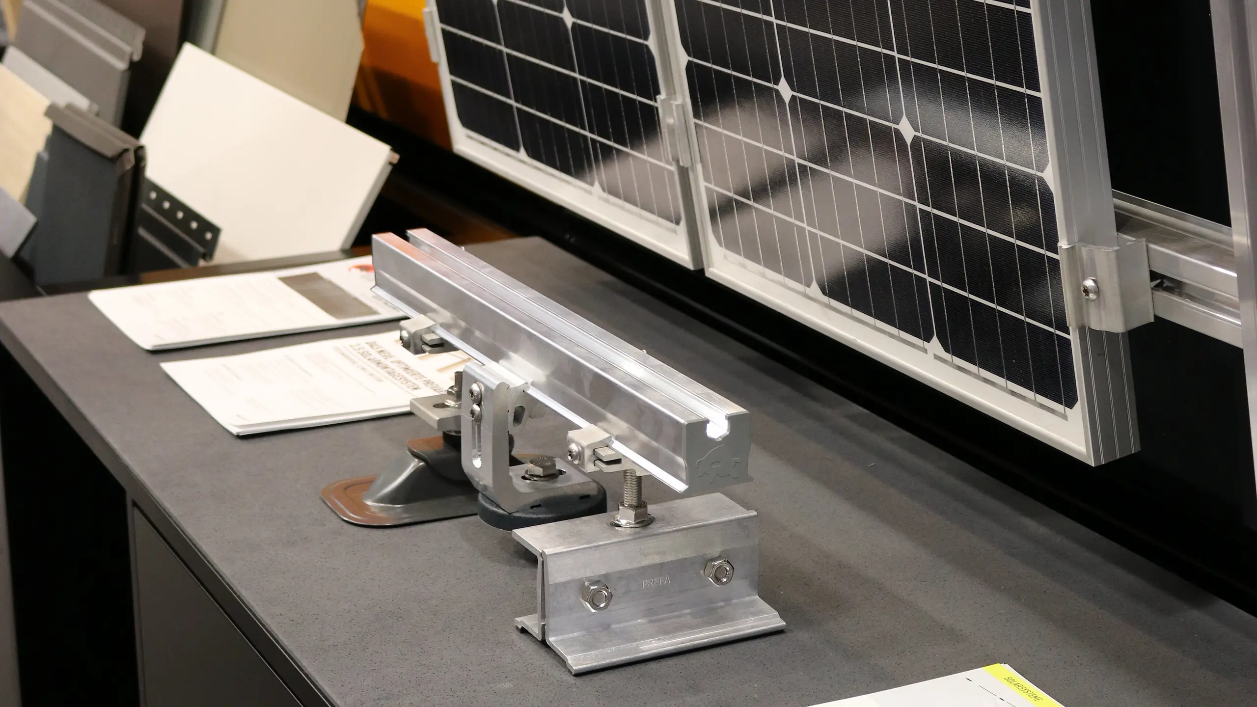 Detailaufnahme des PREFA Messestands mit einem ausgestellten Solarmodul und dem neuen PREVARIO 3.S Solarmontagesystem. Zu sehen sind hochwertige Halterungen und Montageschienen aus Aluminium, die Stabilität und Langlebigkeit garantieren. Daneben liegen technische Informationsblätter zu den Produkten. Im Hintergrund sind Dach- und Fassadenmuster in verschiedenen Farben ausgestellt.