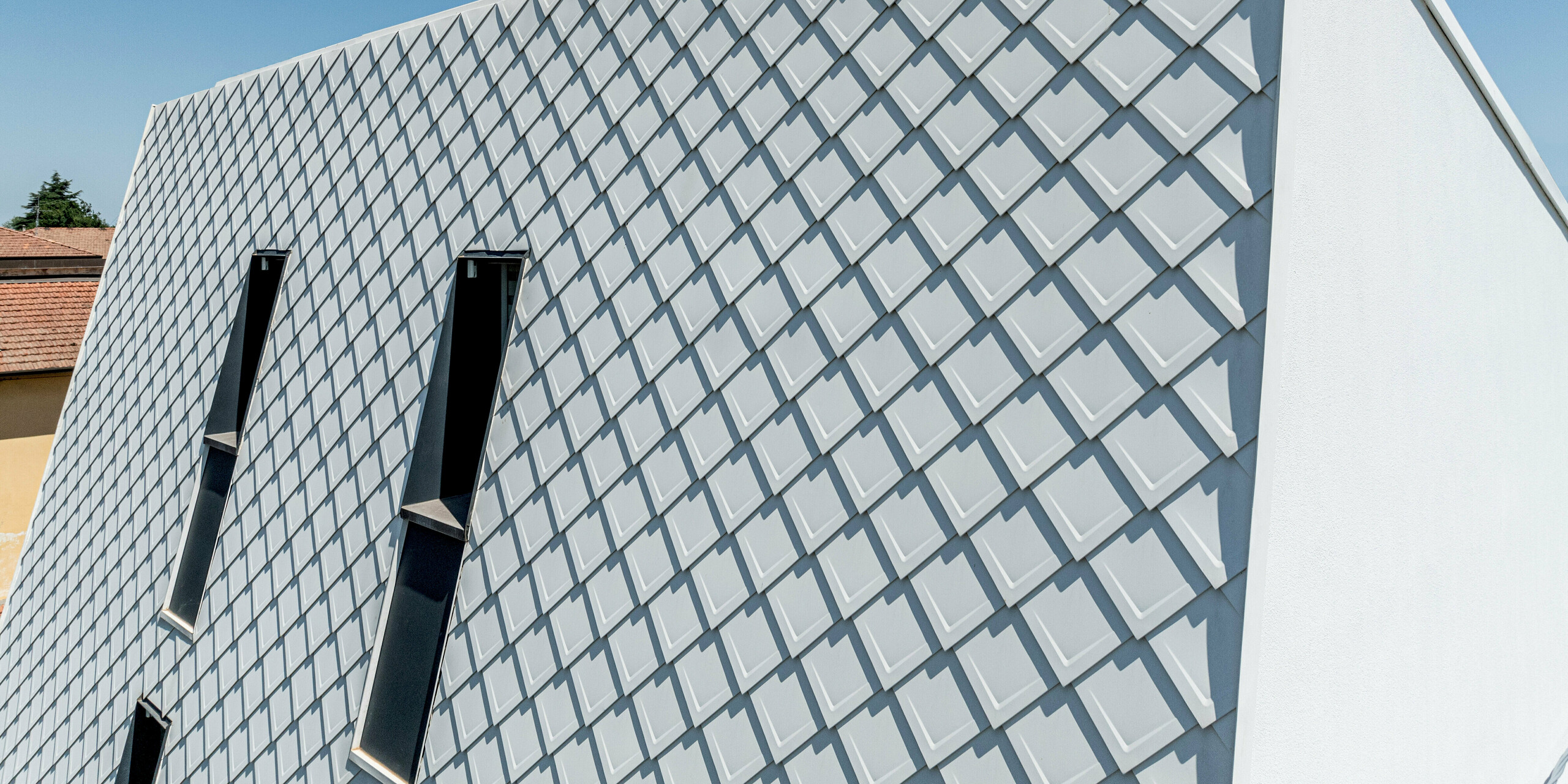 Detailansicht der geneigten Fassadenfläche bis zur Traufe eines modernen Einfamilienhauses in Forlì, Italien. Die PREFA Wandraute 29 × 29 in der Farbe P.10 Reinweiß zieht mit ihrem präzisen, diagonal verlaufenden Aluminiumraster alle Blicke auf sich. Im klaren Tageslicht entstehen markante Licht- und Schattenkanten, die Tiefe und Dynamik erzeugen. Schmale Fensteröffnungen durchbrechen das Rautenmuster gezielt. Die PREFA Aluminiumfassade ist hochwertig, langlebig, witterungsbeständig und überzeugt durch eine dauerhaft makellose Optik.
