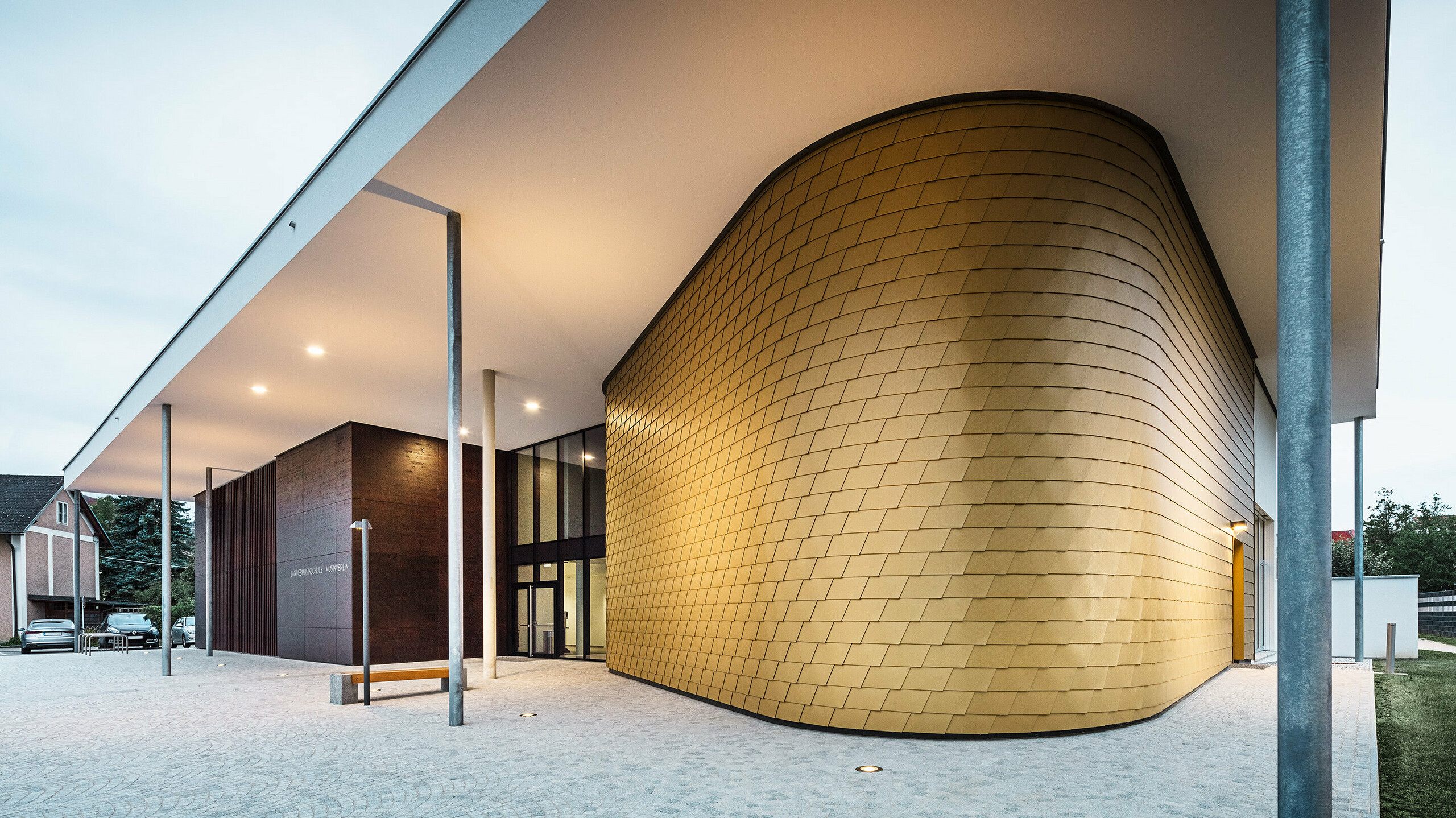 Modernes Gebäude mit markantem Eingangsbereich, dessen geschwungene Fassade mit goldfarbenen Aluminiumrauten verkleidet ist. Das Vordach wird von schlanken Stützen getragen und ist dezent beleuchtet. Neben dem goldenen Fassadenteil befindet sich ein dunkler Kubus mit der Aufschrift „Landesmusikschule Buchkirchen“. Der Vorplatz ist mit hellen Pflastersteinen gestaltet.