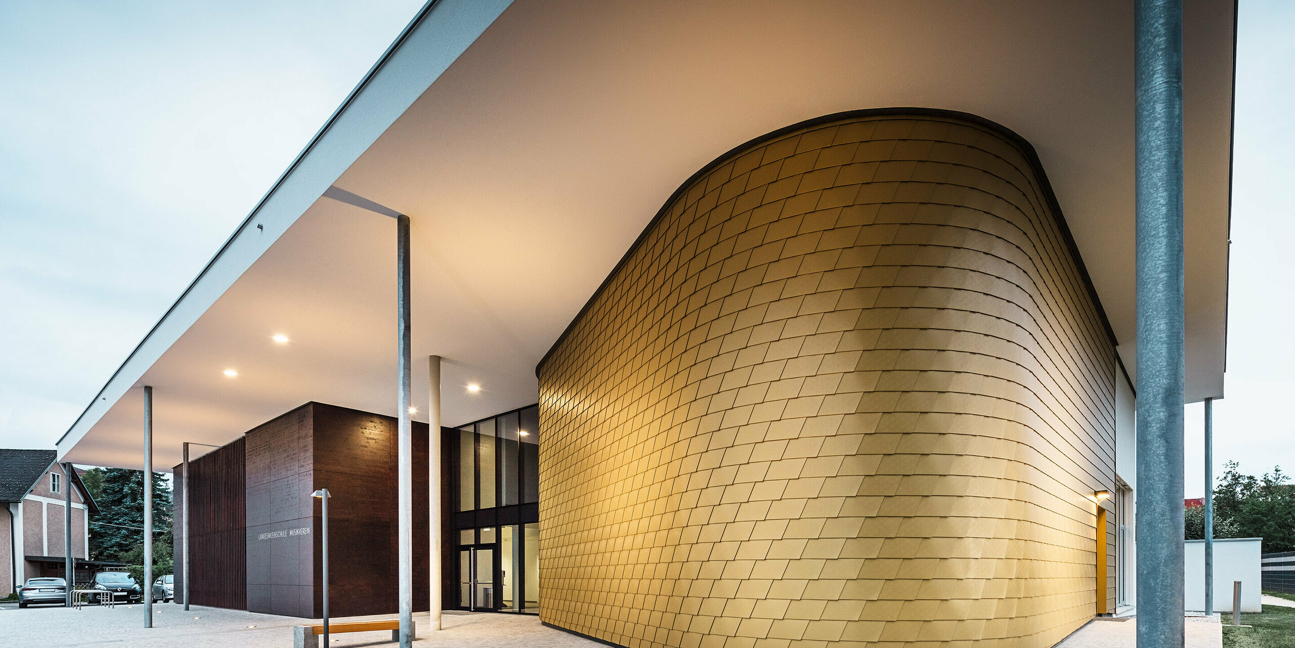 Modernes Gebäude mit markantem Eingangsbereich, dessen geschwungene Fassade mit goldfarbenen Aluminiumrauten verkleidet ist. Das Vordach wird von schlanken Stützen getragen und ist dezent beleuchtet. Neben dem goldenen Fassadenteil befindet sich ein dunkler Kubus mit der Aufschrift „Landesmusikschule Buchkirchen“. Der Vorplatz ist mit hellen Pflastersteinen gestaltet.