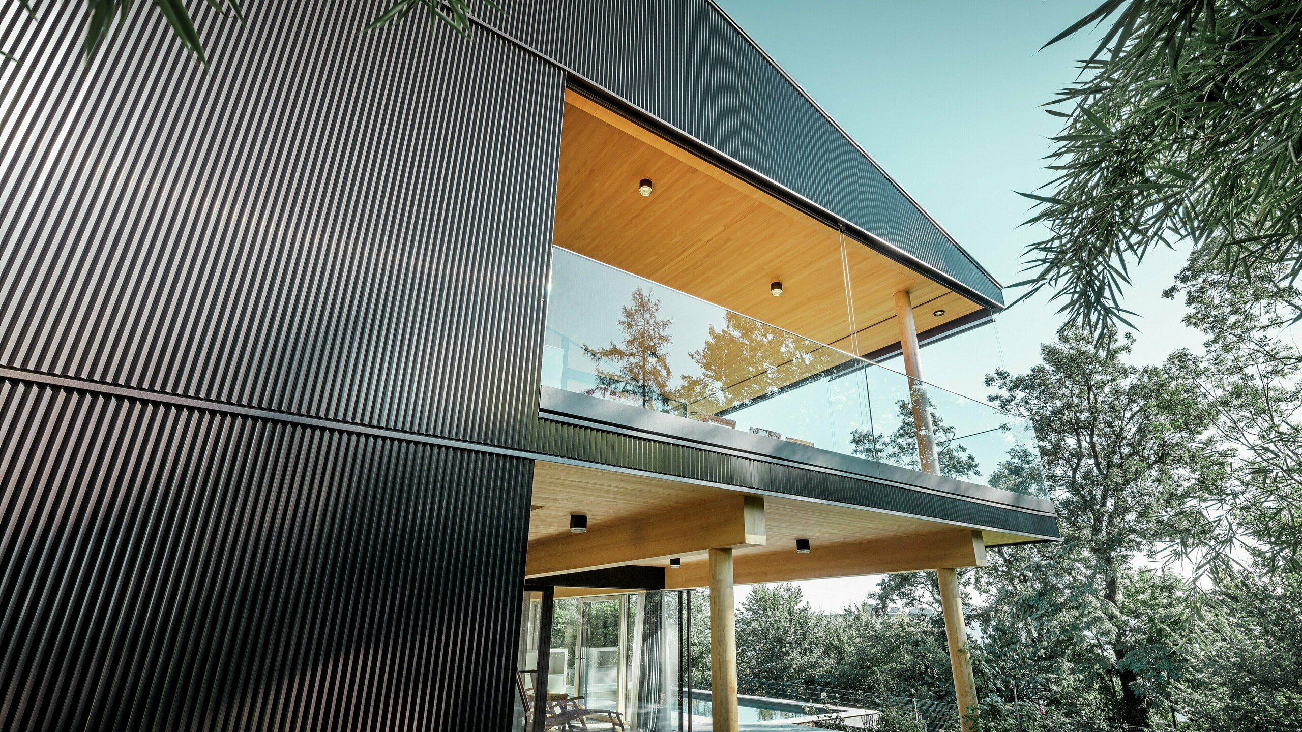 Seitliche Ansicht eines Einfamilienhauses, die Fassade ist mit dunklem PREFA Zackenprofil verkleidet. Man sieht die Terrasse und einen großzügigen Balkon. Die Decke bzw. die Untersicht ist mit Holzpaneelen gestaltet.