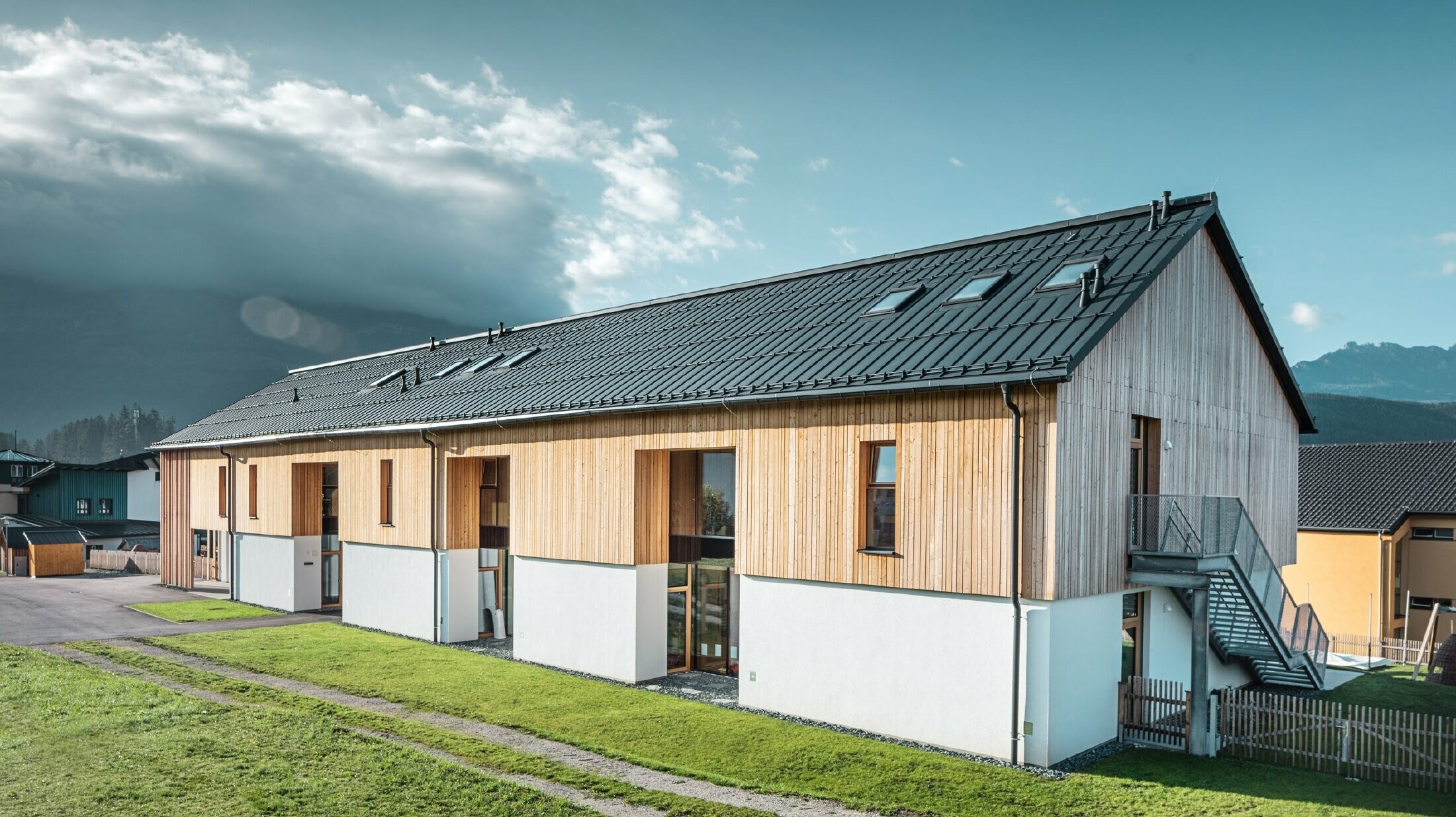 Modernes, langgestrecktes Gebäude mit Satteldach aus dunkelgrauem Aluminium und Fassade aus vertikaler Holzverschalung über weißem Sockel. Große Fensteröffnungen und eine Außentreppe aus Metall betonen die funktionale Architektur. Das Gebäude steht auf einer grünen Wiese mit umliegenden Häusern und Bergen im Hintergrund unter teils wolkigem Himmel.