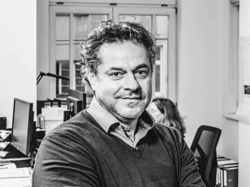 Im Schwarz-Weiß Bild ist der Architekt Ralf Mühlbacher auf einem Tisch sitzend zu sehen.