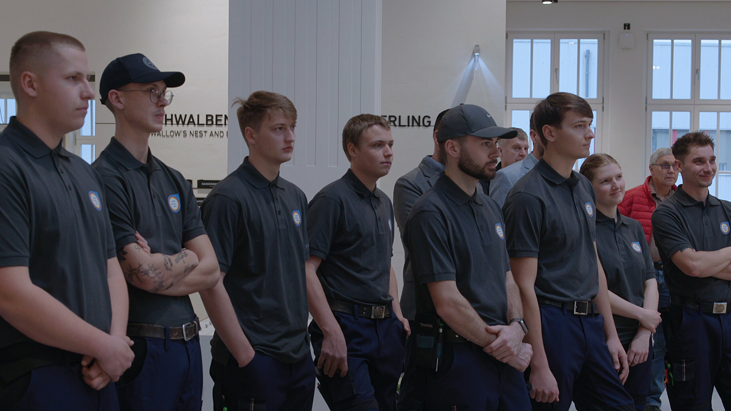 Die Gruppe junger Klempnerinnen und Klempner hört gespannt zu, während sie sich der anspruchsvollen Aufgabe der Deutschen Meisterschaft im Klempnerhandwerk stellen. Der Raum ist modern und lichtdurchflutet, im Hintergrund sind Zuschauer und Branchenvertreter zu sehen.