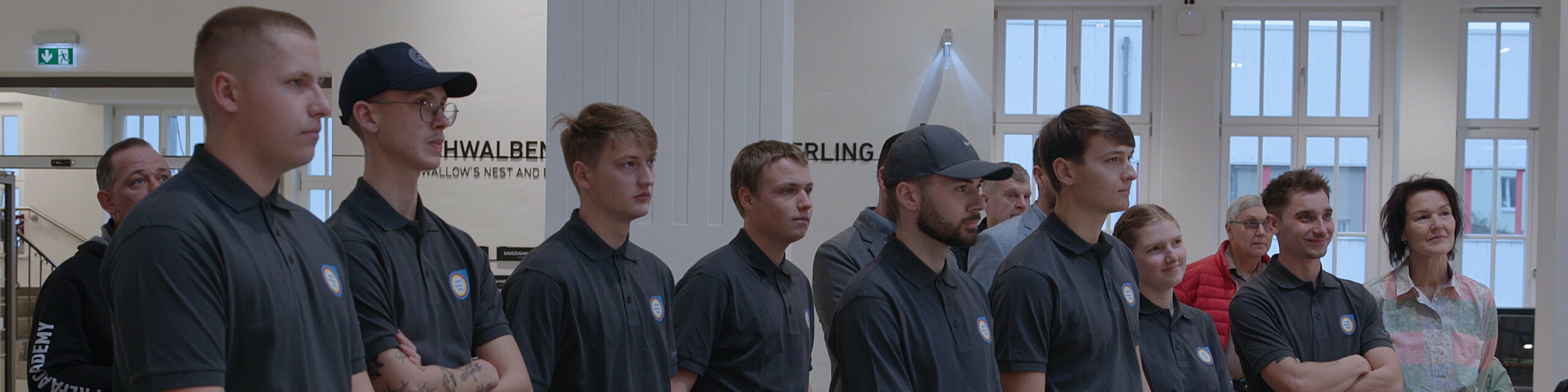 Die Gruppe junger Klempnerinnen und Klempner hört gespannt zu, während sie sich der anspruchsvollen Aufgabe der Deutschen Meisterschaft im Klempnerhandwerk stellen. Der Raum ist modern und lichtdurchflutet, im Hintergrund sind Zuschauer und Branchenvertreter zu sehen.