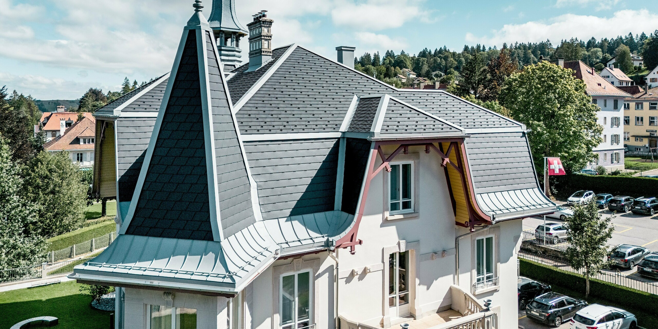 Faszinierende Detailansicht der Manufacture Horlogère du Locle in der Schweiz mit markantem Turm als architektonischem Highlight. Die vielschichtige Dachlandschaft beeindruckt durch steil geneigte Flächen und elegante Rundungen. PREFA Dachschindeln in P.10 Schwarz verleihen der Struktur klare Linien und ein dynamisches Relief, während PREFALZ in Patinagrau geschwungene Übergänge und komplexe Formen präzise umsetzt. Das leichte Aluminiumdach überzeugt durch Langlebigkeit, hohe Witterungsbeständigkeit und maximale Gestaltungsfreiheit bei anspruchsvoller Dacharchitektur.