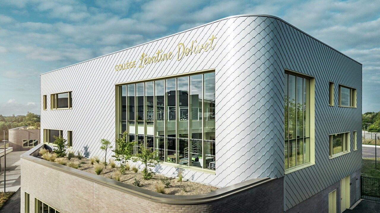 Das Foto zeigt eine markante Ecke des Collège Léontine Dolivet in Cesson-Sévigné. Die Fassade besteht aus silbermetallischen PREFA-Wandelementen, die diagonal angeordnet sind und das Licht reflektieren. Besonders auffällig ist die geschwungene Form des Obergeschosses, die dem Gebäude eine sanfte und moderne Anmutung verleiht. Große Glasflächen geben den Blick ins Innere frei, während gelbe Fensterrahmen farbliche Akzente setzen. Im Vordergrund befindet sich eine bepflanzte Fläche mit Kies und Sträuchern, die die Architektur harmonisch ergänzt. Der leicht bewölkte Himmel hebt die Metalloberfläche zusätzlich hervor.