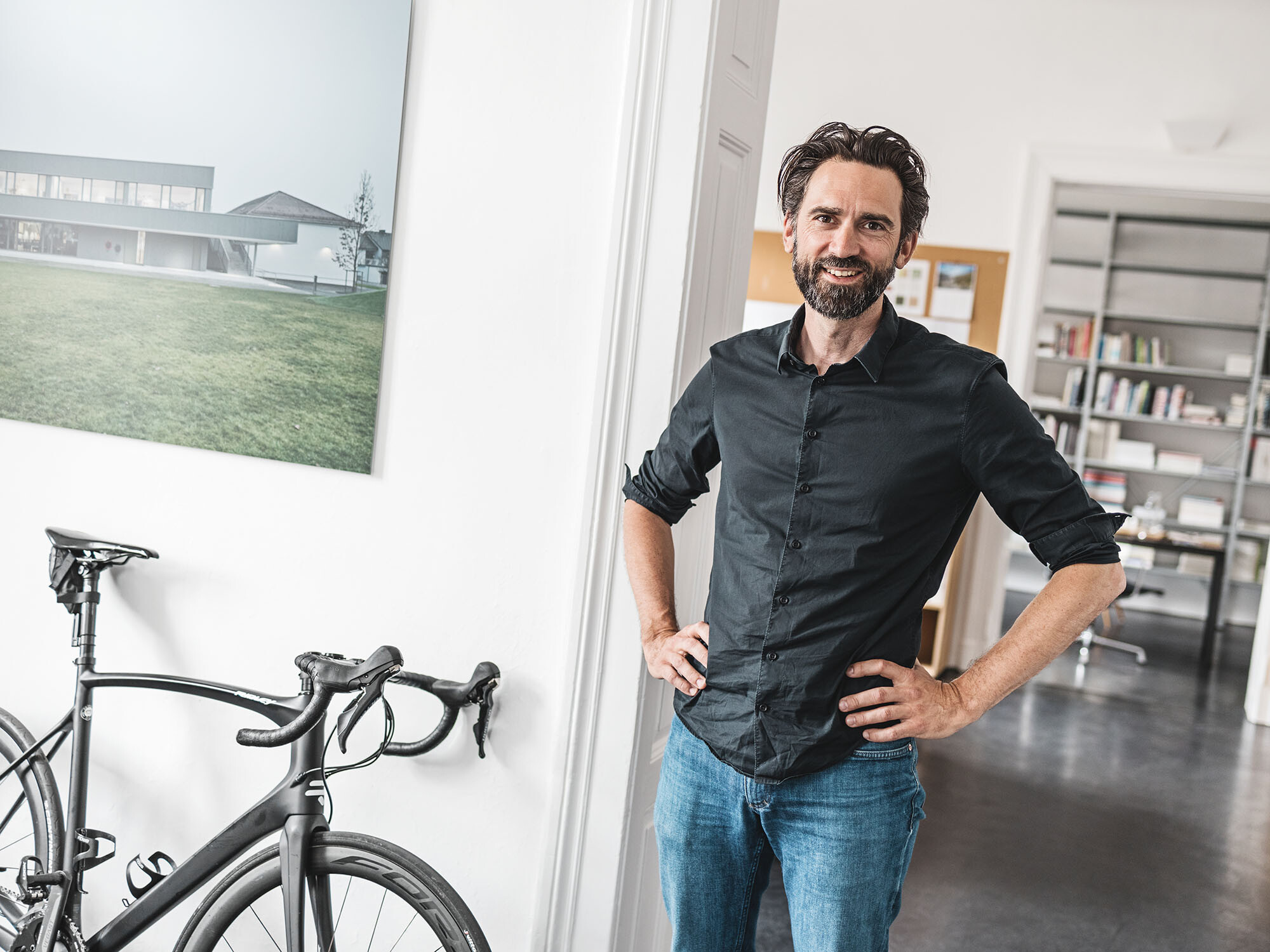 Portrait des leitenden Architekten Thomas Heil; hinter ihm erstreckt sich ein Büroraum, neben ihm ist ein Fahrrad an die Wand gelehnt.