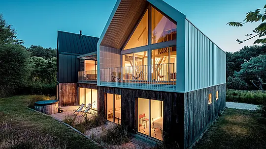 Das moderne Wohnensemble Le Rhune beeindruckt in der blauen Stunde mit seiner markanten Architektur aus zwei versetzt angeordneten Baukörpern. Die helle PREFALZ Aluminiumhülle umschließt Dach und Fassade des Vorderhauses als präzise geformte Einheit und betont die klare Geometrie des Satteldachs. Großzügige Glasflächen lassen warmes Licht nach außen strahlen und schaffen einen faszinierenden Kontrast zur umgebenden Natur. Die langlebigen und wetterbeständigen PREFALZ Stehfalzsysteme ermöglichen filigrane Details, eine durchgängige Gebäudehülle sowie eine zeitgemäße Interpretation traditioneller bretonischer Bauformen.