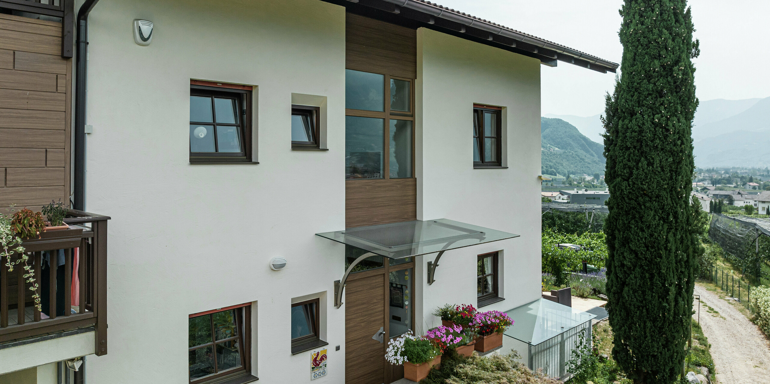 Eingangsbereich der Ferienwohnungen Prechtlhof in Meran, Südtirol, mit moderner Fassadengestaltung aus Putz und PREFA Sidings in Walnussbraun. Die wartungsfreie Aluminiumfassade in Holzoptik ergänzt das traditionelle Design und sorgt für eine langlebige und witterungsbeständige Lösung.
