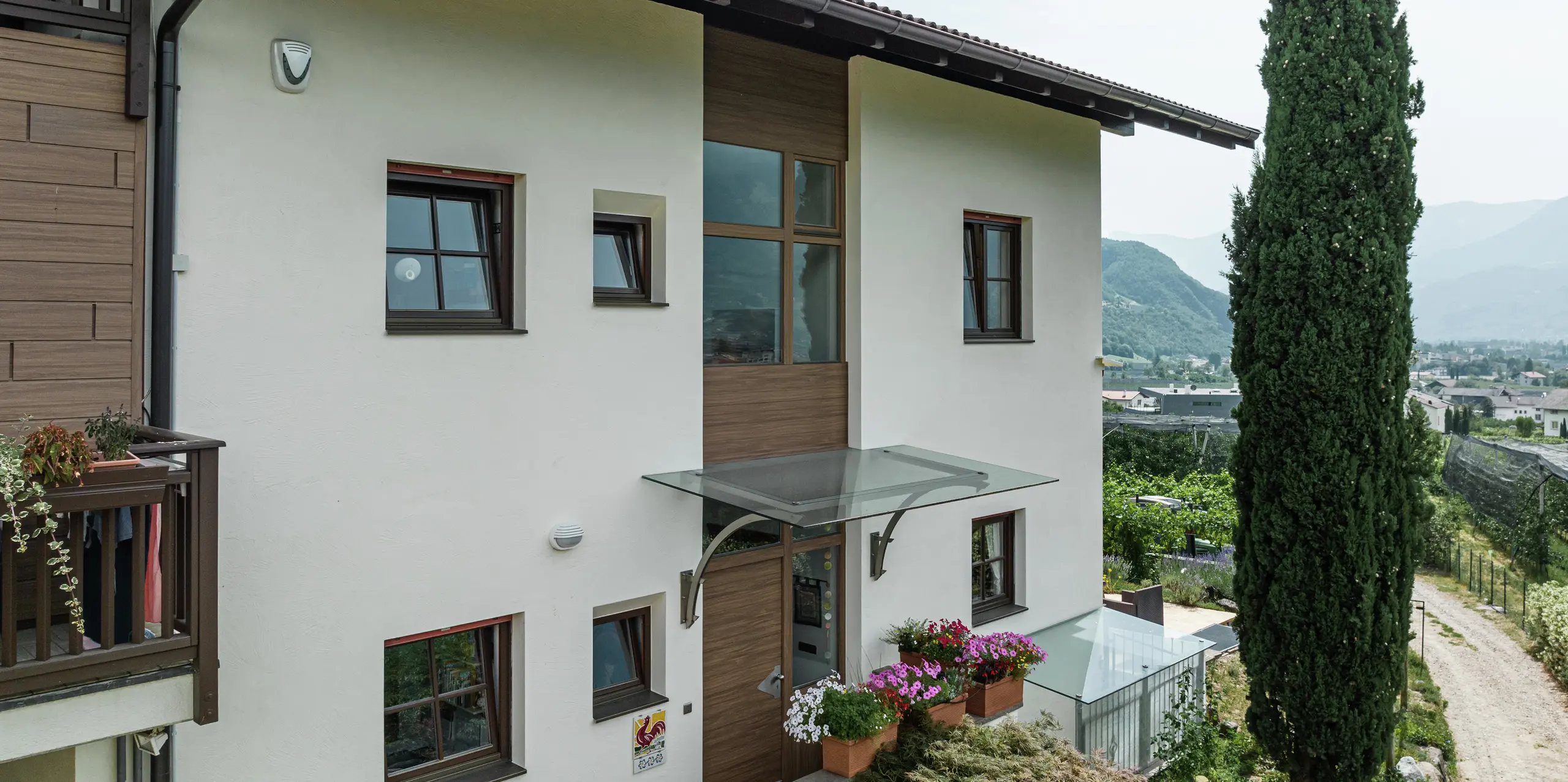 Eingangsbereich der Ferienwohnungen Prechtlhof in Meran, Südtirol, mit moderner Fassadengestaltung aus Putz und PREFA Sidings in Walnussbraun. Die wartungsfreie Aluminiumfassade in Holzoptik ergänzt das traditionelle Design und sorgt für eine langlebige und witterungsbeständige Lösung.