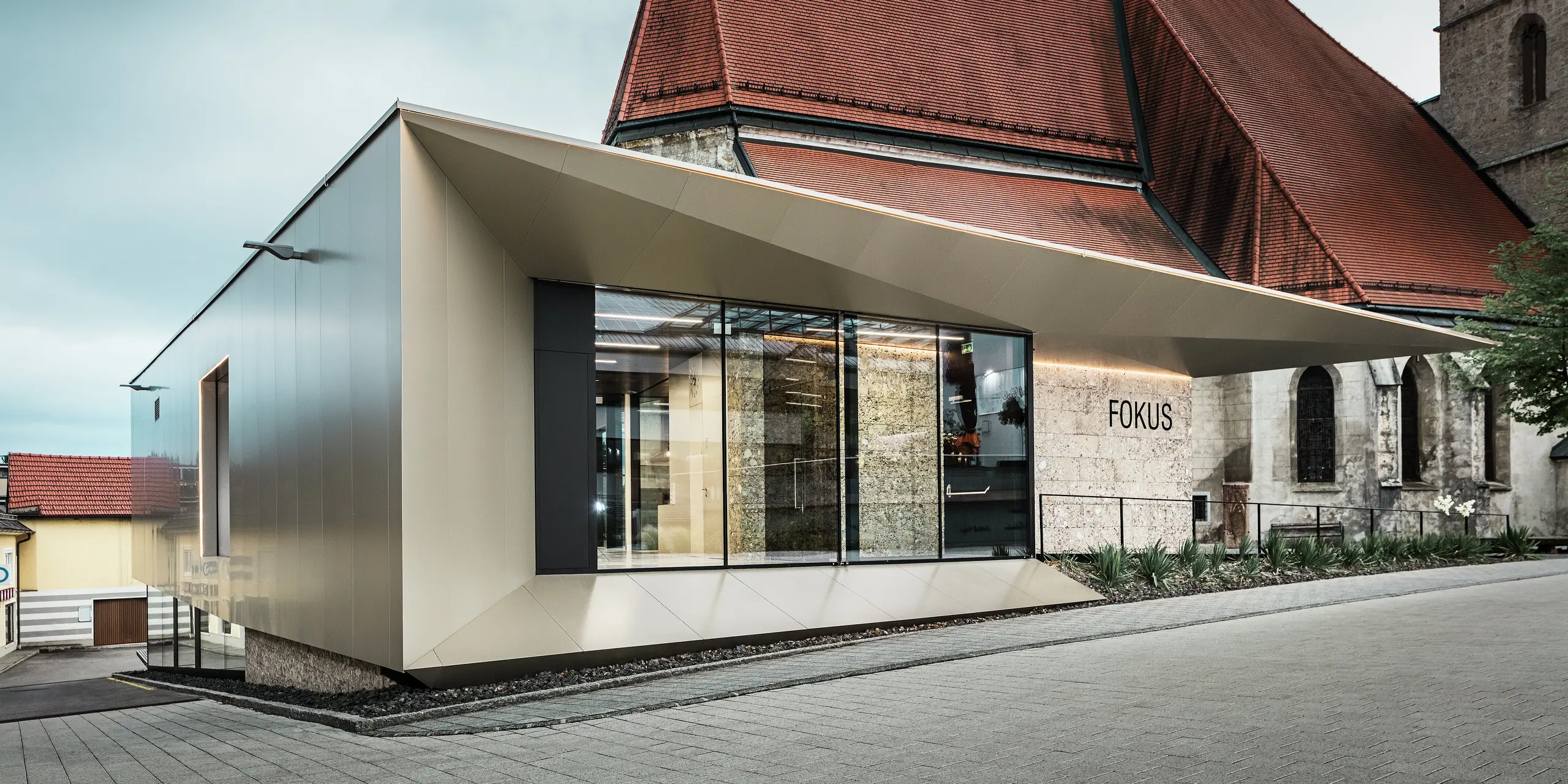 Im Vordergrund des Pfarrheims in Sierning glänzt die Aluminiumfassade durch ihre saubere Linienführung und hohe Formstabilität. Die Kombination aus Glasflächen und bronzefarbenen PREFA Aluminium Verbundplatten betont Offenheit, Beständigkeit und gestalterische Präzision bei gleichzeitiger Integration ins historische Umfeld.