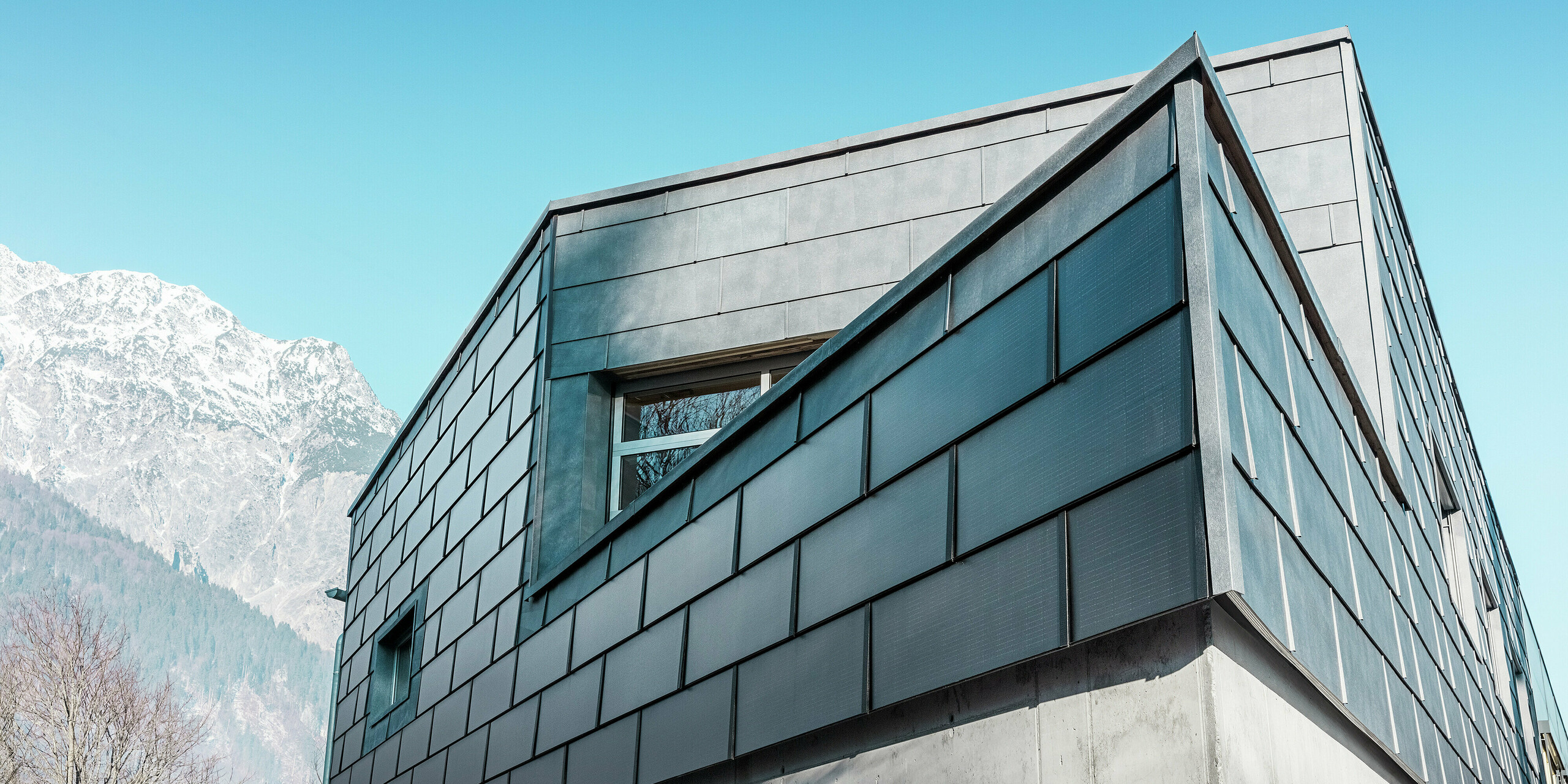 Fassade des Firmengebäudes der Spenglerei RÜMA in Vorarlberg mit eleganten, grauen PREFA Solardachplatten und PREFA Fassadenpaneelen FX.12, kombiniert mit Sichtbeton, vor klar blauem Himmel und Bergkulisse. Die spitze Gebäudeecke unterstreicht die auffallende Architektur.