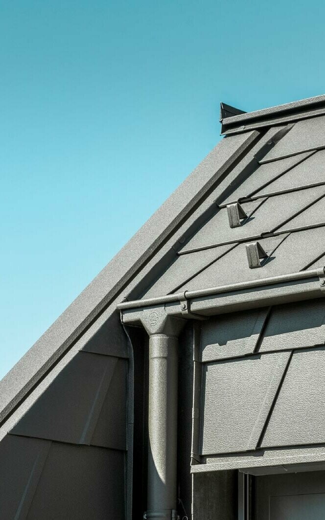Dach mit PREFA Aluminiumdachplatten und modernen Regenrinnen, die eine hohe Langlebigkeit und ästhetische Funktionalität bieten, vor einem klaren blauen Himmel.