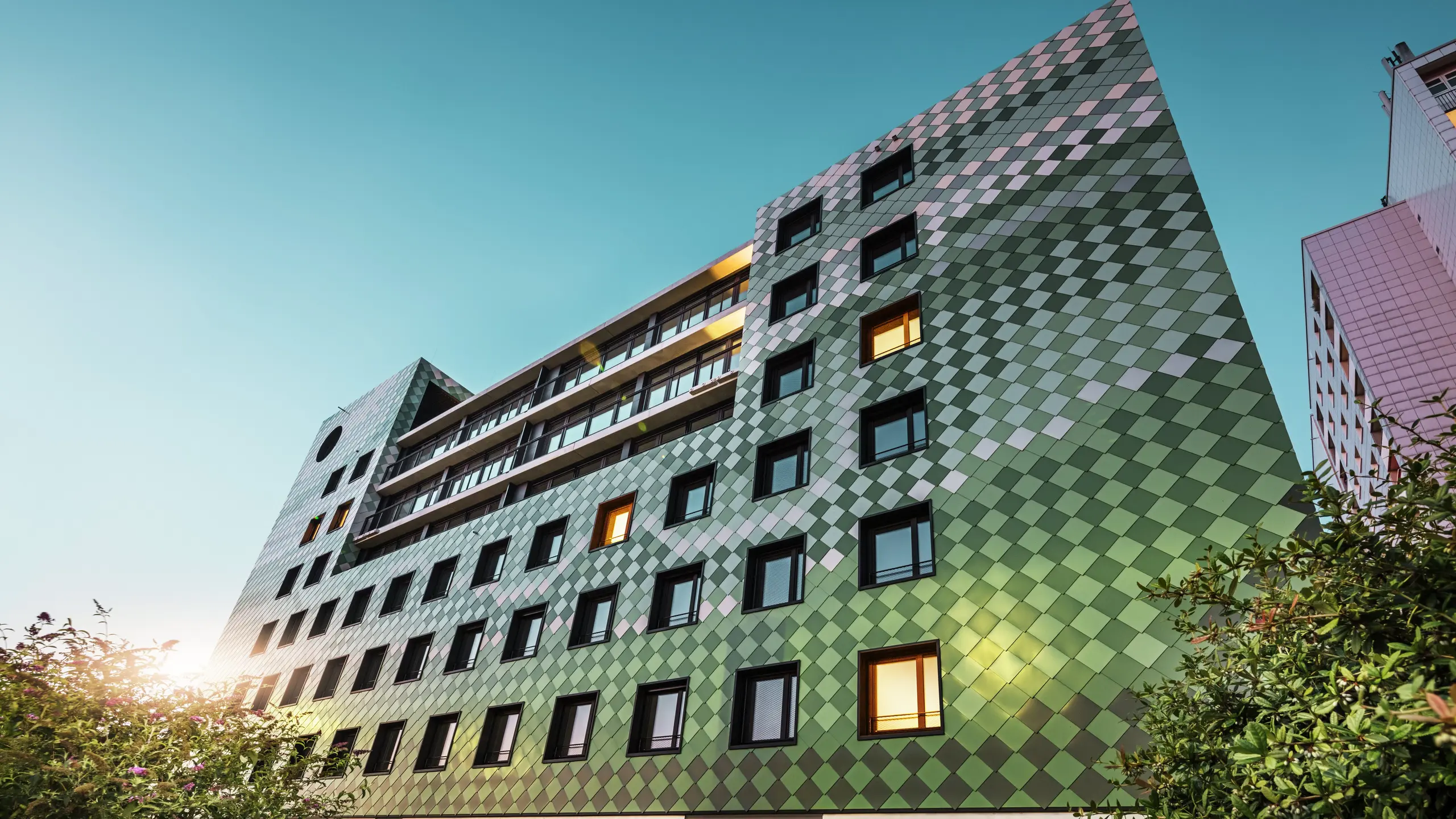 Das moderne Wohn- und Geschäftsgebäude „Pick-Nick“ in Dresden besticht durch seine markante PREFA-Fassade aus Aluminium-Rhombusen in den Sonderfarben Olivgrün, Graugrün, Resedagrün, Opalgrün, Hellelfenbein, Perlweiß und Hellbronze. Die kreative Fassadengestaltung erinnert an ein Reisfeld und erzeugt ein lebendiges Farbenspiel. Die klare Architektur, die schwarzen Fensterrahmen und die Langlebigkeit der PREFA-Materialien prägen das Gebäude. Das tiefstehende Sonnenlicht spiegelt sich auf der Fassade und den Fensterflächen.
