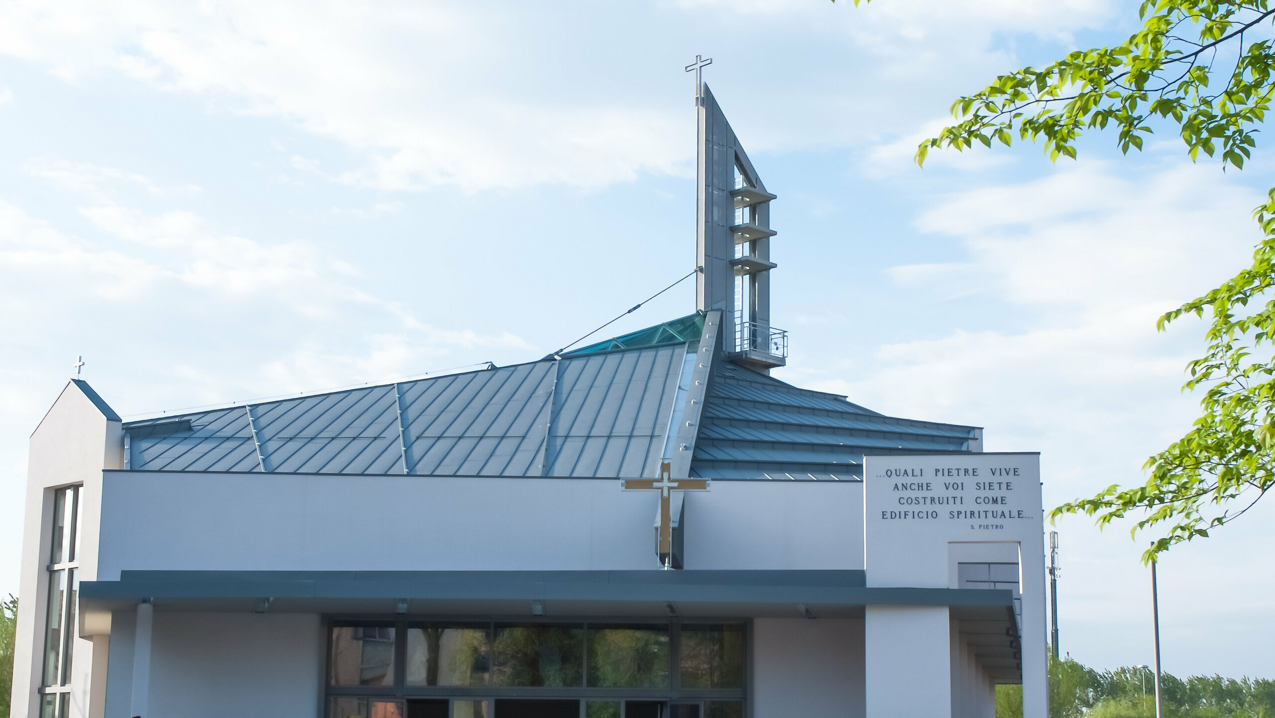 Die Kirche Parrocchia San Lazzaro ist mit PREFALZ in P.10 Hellgrau eingedeckt. Das moderne Kirchengebäude hat ein schräges Dach und eine charakteristische, geometrische Architektur. Auf dem Vorplatz der Kirche stehen einige Personen, die sich unterhalten. Die Fassade ist schlicht gestaltet, mit großen Fenstern und einem hervorgehobenen Eingang. Rechts an der Wand befindet sich eine Inschrift auf Italienisch. 