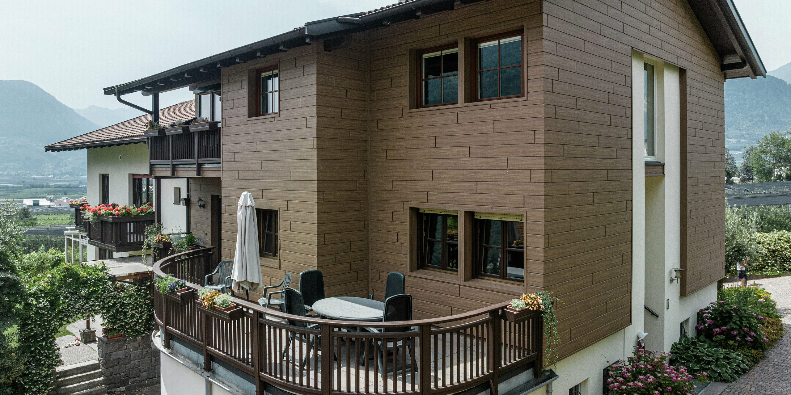 Sanierte Ferienwohnungen Prechtlhof in Meran, Südtirol, mit PREFA Sidings in Walnussbraun. Die wartungsfreie Aluminiumfassade in Holzoptik verbindet moderne Ästhetik mit traditionellem Charme und fügt sich harmonisch in die alpine Umgebung ein. Der Rundbalkon im Vordergrund lädt zum Verweilen in der traumhaften Südtiroler Landschaft ein. 