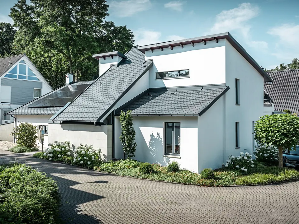 Modernes Einfamilienhaus mit weißer Putzfassade und dunklem PREFA Schrägdach aus Aluminium. Auf dem Dach sind integrierte Photovoltaikmodule und eine Aluminium-Dachentwässerung in Anthrazit zu sehen. Das Gebäude liegt in einer gepflegten Wohnsiedlung mit bepflanzten Vorgärten, gepflasterter Zufahrt und umliegenden Einfamilienhäusern.