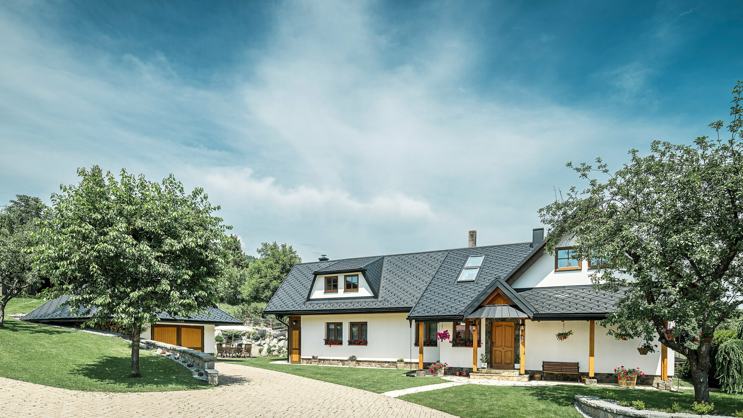 Zufahrt und Eingang eines traditionellen Einfamilienhauses in Stara Bystrica, Slowakei. Das Gebäude ist mit PREFA Dachrauten 29 × 29 in P.10 Schwarz gedeckt. Die Aluminiumrauten sorgen für robusten und langlebigen Schutz. Die gepflegte Pflastereinfahrt und die umgebende grüne Landschaft, mit einem Baum im Vordergrund, ergänzen das harmonische Gesamtbild des Hauses.
