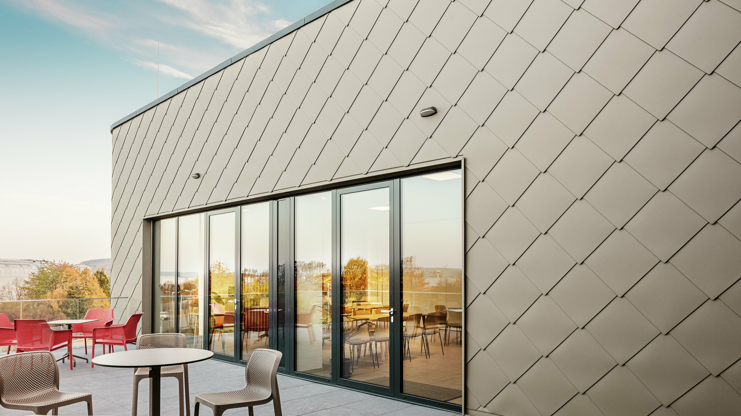 Die Fassade des neuen PREFA Standortes in Törökbálint besticht durch die eleganten PREFA Wandrauten 44 × 44 in Bronze. Die präzise verlegte Aluminiumfassade sorgt für ein modernes, dynamisches Erscheinungsbild und bietet gleichzeitig maximalen Schutz vor Witterungseinflüssen. Die großzügige Glasfront verbindet Innen- und Außenbereich und schafft auf der großzügigen Terrasse eine helle und einladende Atmosphäre.