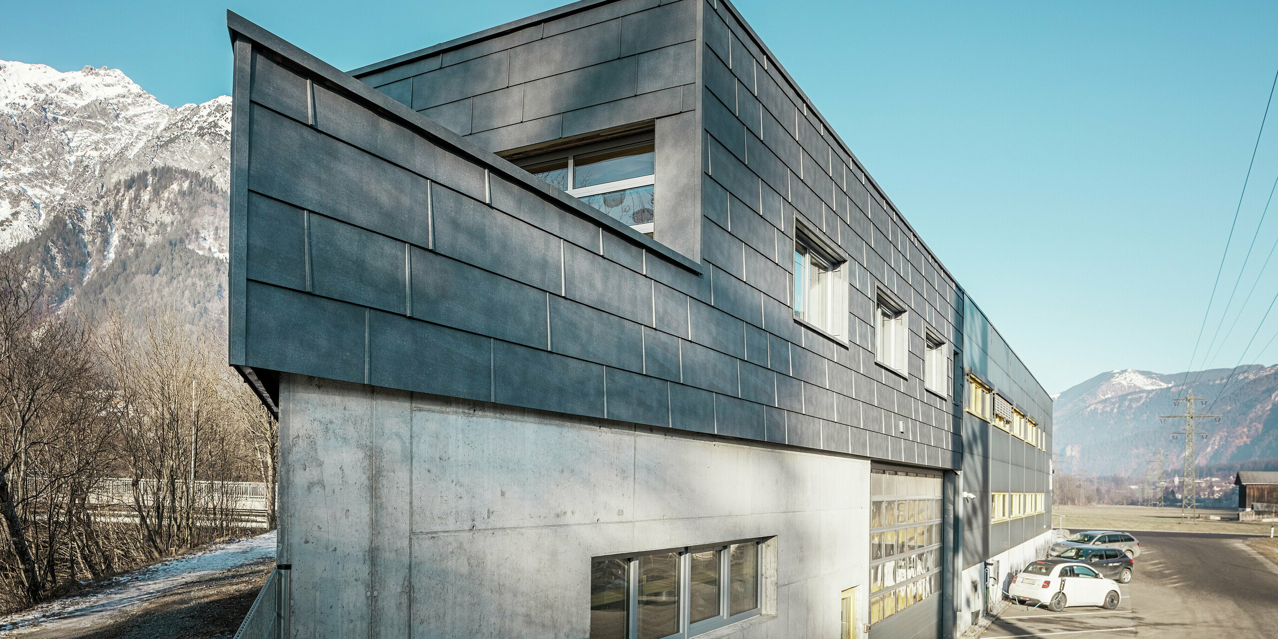 Die Vorderseite des Firmengebäudes der Spenglerei RÜMA in Vorarlberg. Es verfügt über eine markante Fassade aus grauen PREFA FX.12-Fassadenpaneelen aus Aluminium und einen sichtbaren Betonunterbau. Es ist in eine alpine Landschaft mit Bergen und blauem Himmel eingebettet. Auf dem Parkplatz vor dem Gebäude ist ein ladendes Elektroauto zu sehen, das die Nachhaltigkeit des Unternehmens unterstreicht.