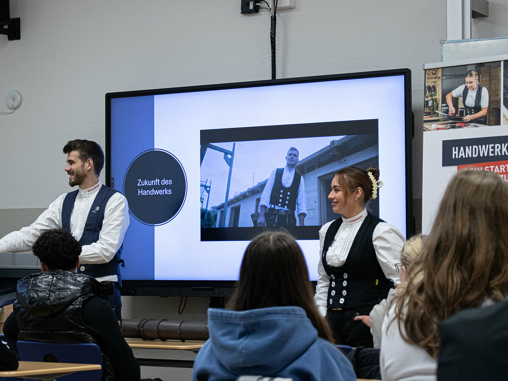 Klempnermeister Marvin Bürger und Dachdeckermeisterin Fabienne Ellermeier präsentieren den Schülerinnen und Schülern während der Handwerker-Tour 2025 eine Präsentation zum Thema ‚Zukunft des Handwerks‘. Auf dem Bildschirm hinter ihnen ist ein Bild eines Handwerkers zu sehen. Fabienne Ellermeier steht neben Marvin Bürger und lächelt, während die Schülerinnen und Schüler aufmerksam zuhören.