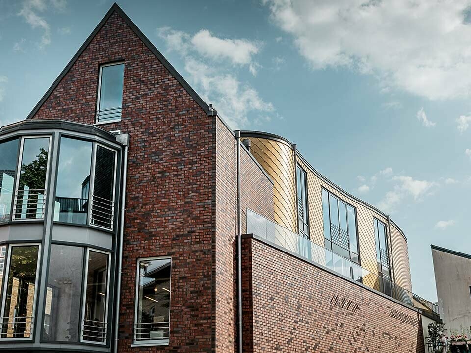 neues Schuhhaus Schüttfort in Hamburg-Bergedorf mit einer klassischen Klinkerfassade und einem geschwungenen Fassadenteil, verkleidet mit einer Aluraute von PREFA, das Satteldach ist in grau mit Aluminium gedeckt, viele Glas- und Fensterflächen, leicht bewölkt