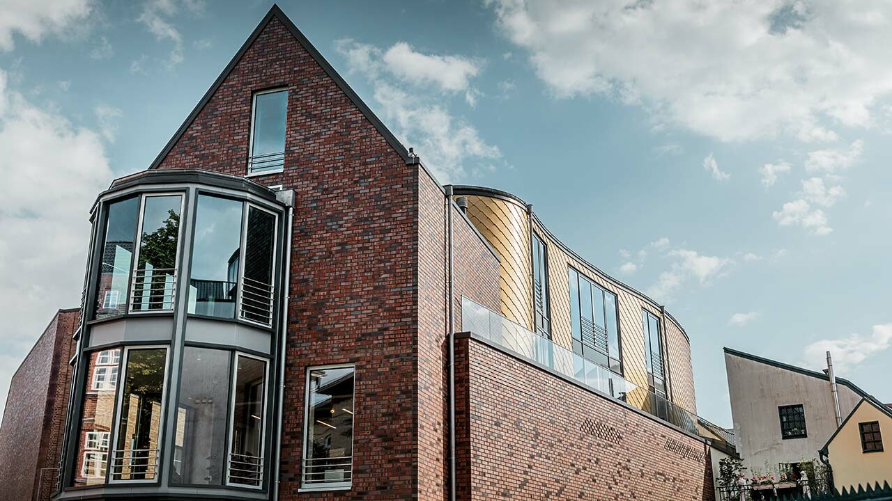 neues Schuhhaus Schüttfort in Hamburg-Bergedorf mit einer klassischen Klinkerfassade und einem geschwungenen Fassadenteil, verkleidet mit einer Aluraute von PREFA, das Satteldach ist in grau mit Aluminium gedeckt, viele Glas- und Fensterflächen, leicht bewölkt