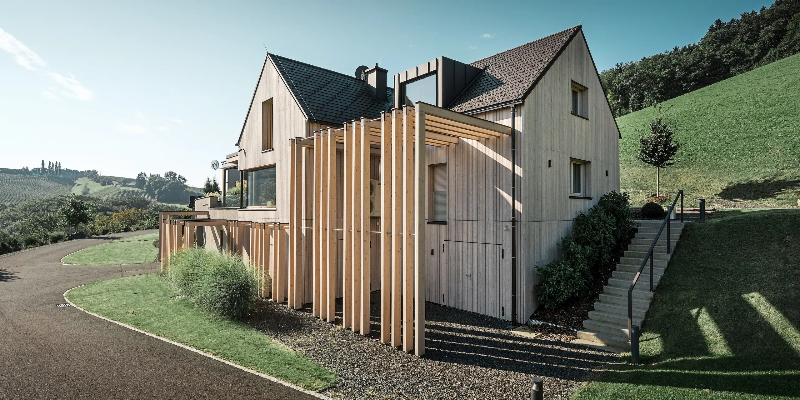 Großes Einfamilienhaus mit Satteldach und Gaube in Hanglage. Das Dach ist mit braunen PREFA Aluminium Dachschindeln eingedeckt und die Fassade mit vertikalen Holzbrettern verkleidet.