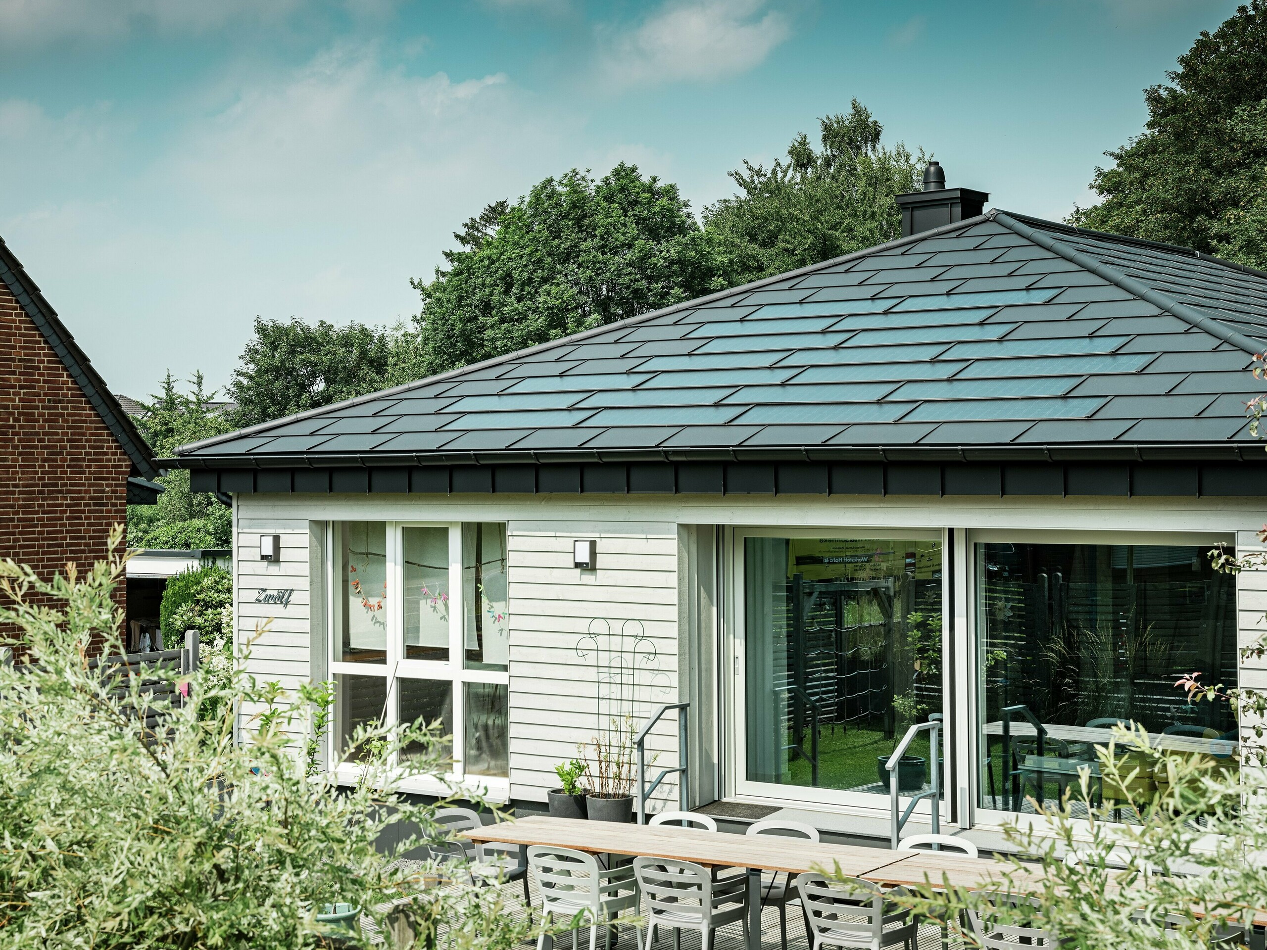 Sanierter Bungalow in Rommerskirchen mit elegantem PREFA Aluminiumdach. PREFA Solardachplatten und Dachplatten R.16 in P.10 Schwarz schützen dauerhaft und erzeugen nachhaltig Strom aus Sonnenenergie. Die großzügige Fensterfront sorgt für viel Licht, die Dachentwässerung mit Saumrinne und Ablaufrohr verbindet Funktionalität und Design perfekt.
