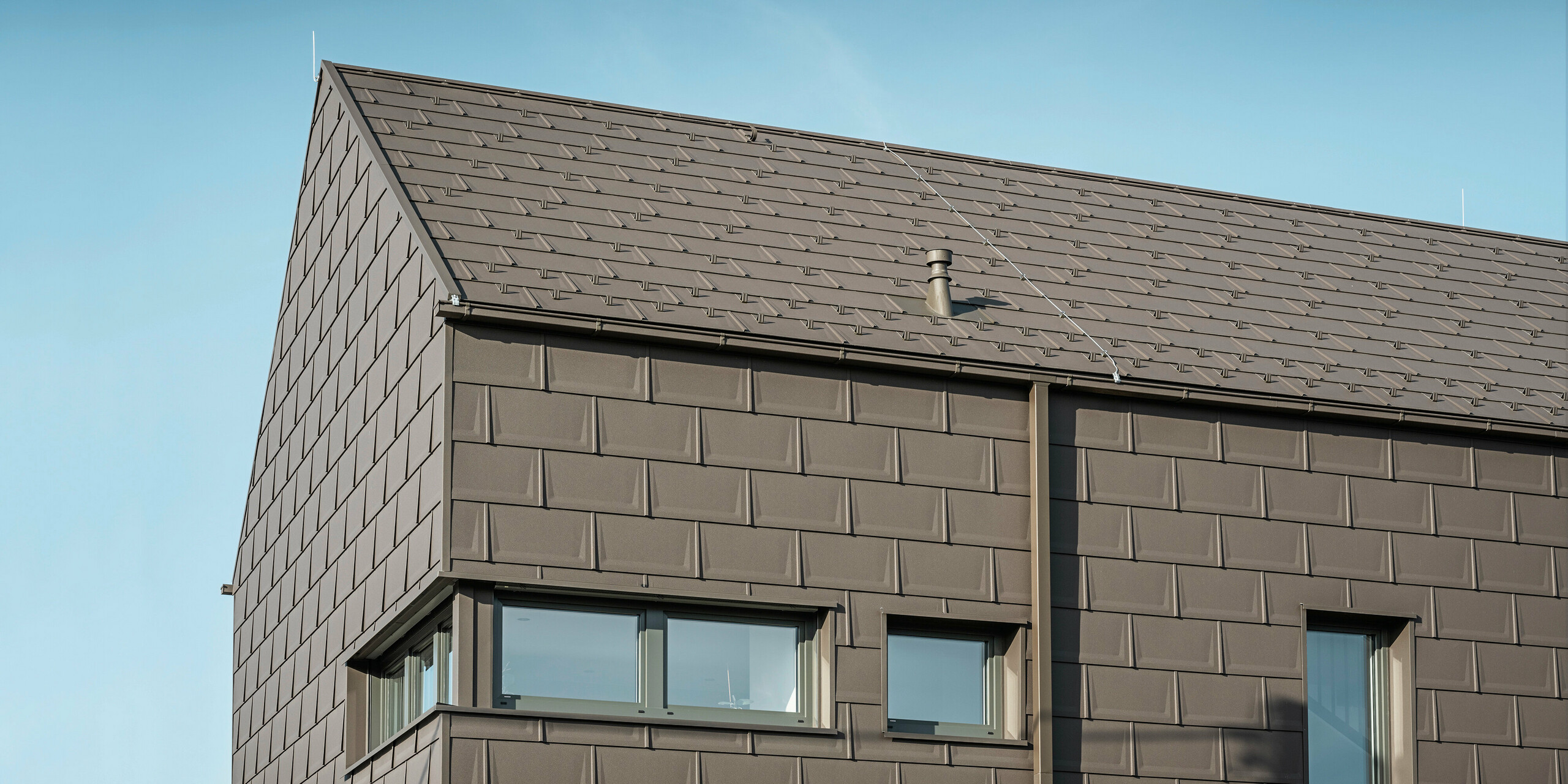 Detailansicht eines Einfamilienhauses in Neukirchen, Österreich, mit einer Dach- und Fassadenverkleidung aus PREFA Dachplatten R.16 in P.10 Braun. Die präzise Überlappung der Platten sorgt für ein einheitliches und ansprechendes Erscheinungsbild, das moderne Ästhetik mit Funktionalität verbindet. Die integrierte PREFA Dachentwässerung in Form der Kastenrinne und des Quadratrohrs in P.10 Braun zeigt das Engagement von PREFA für qualitativ hochwertige und optisch ansprechende Baukomponenten.