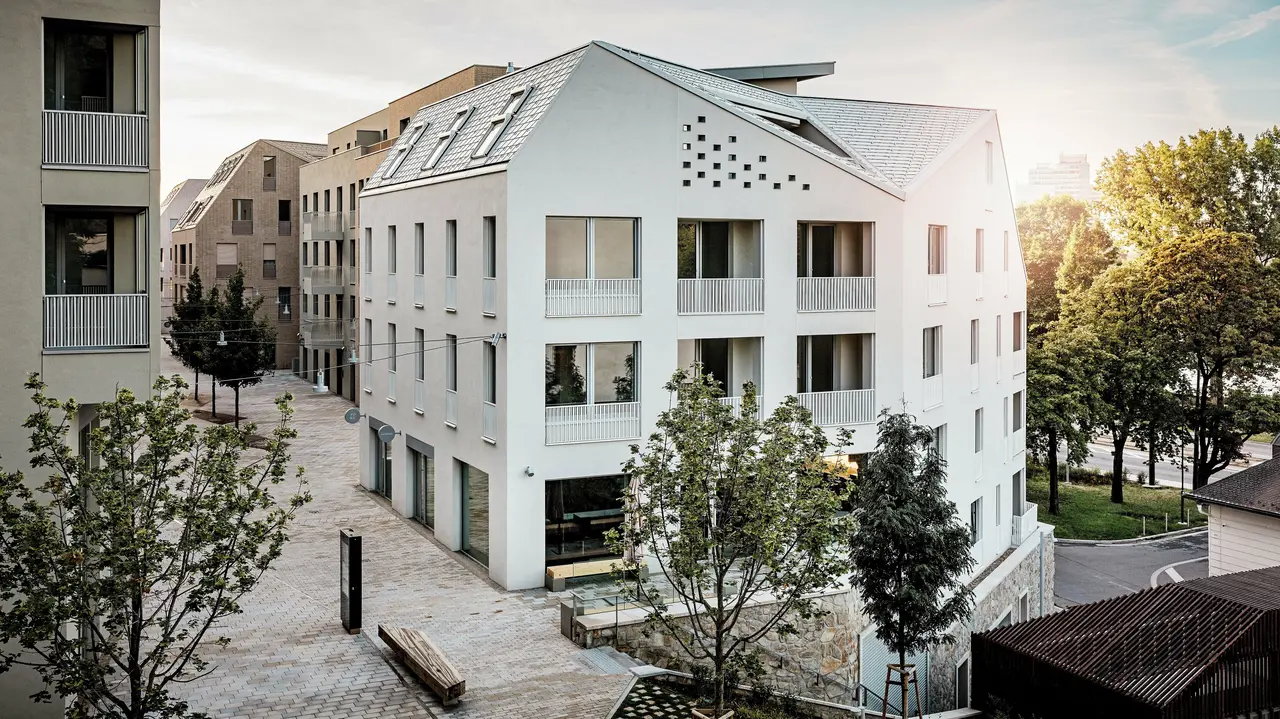 Modernes Wohnquartier Vydrica in Bratislava mit klarer Architektursprache und ruhiger Gassenatmosphäre: Die steil geneigten Satteldächer mit markanten Dachfenstern sind mit PREFA Aluminium Dachschindeln in P.10 Prefaweiß, Bronze und P.10 Sandbraun gedeckt und bilden einen eleganten Eyecatcher im Sonnenlicht. Die präzise ausgeführten Linien und homogenen Flächen unterstreichen die hochwertige Gestaltung. PREFA Aluminium überzeugt durch Langlebigkeit, geringes Gewicht und hohe Witterungsbeständigkeit – ideal für komplexe Dachformen und eine nachhaltige, wartungsarme Gebäudehülle.