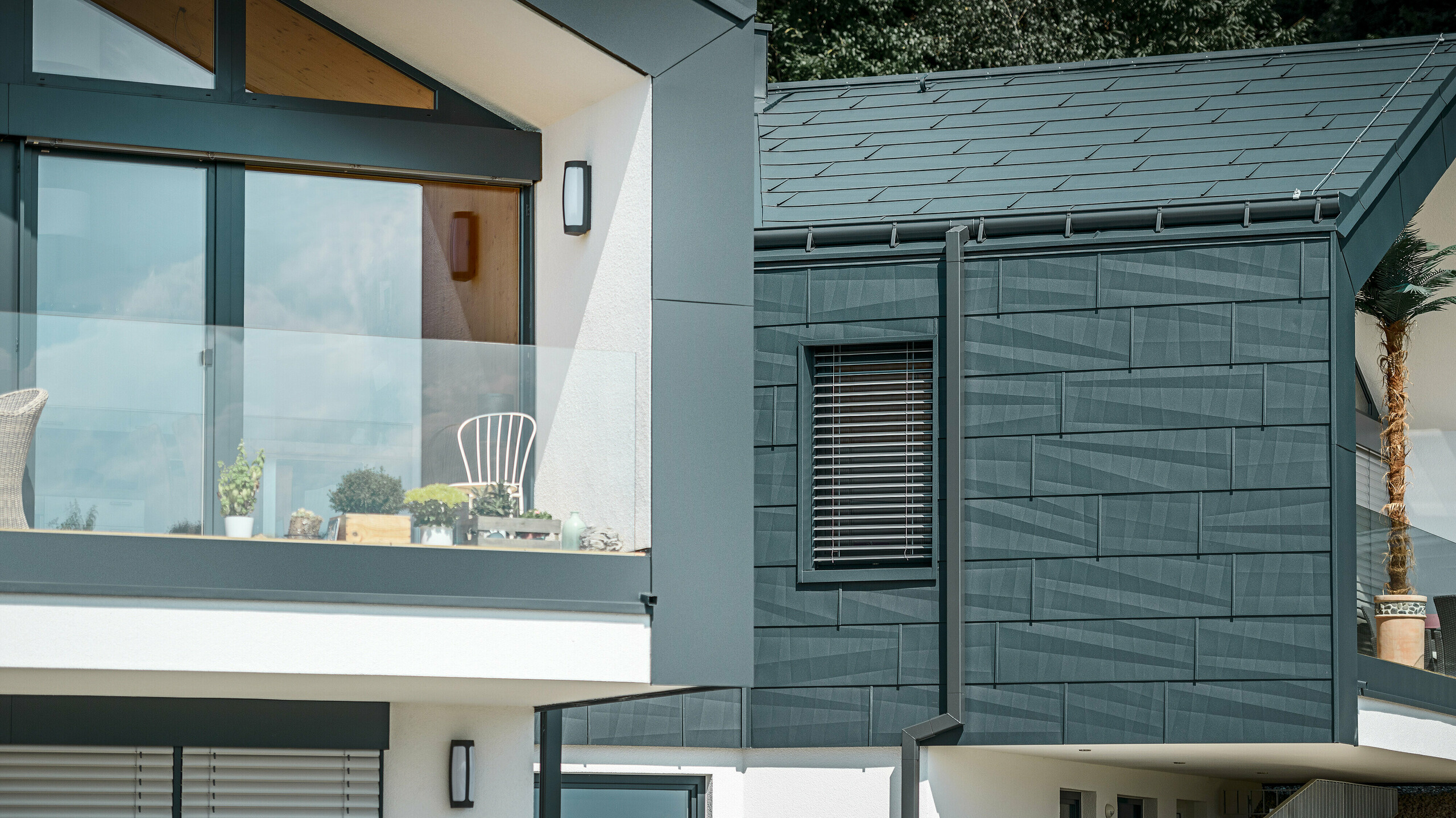 Das Bild zeigt ein modernes Doppelhaus mit einer stilvollen Fassade aus Aluminium. Die Fassade ist in einem dunklen Anthrazitton gehalten und hat eine strukturierte Oberfläche durch das PREFA Dach- und Fassadenpaneel FX.12. Am Dach wurde eine Saumrinne und das Quadratrohr als Ablaufrohr von PREFA montiert. Ein Balkon mit Glasgeländer ist an der oberen Etage zu sehen, während an der unteren Etage Fenster mit Lamellenjalousien zu finden sind. Die Architektur wirkt klar und minimalistisch, und im Hintergrund sind Bäume zu erkennen, die eine natürliche Atmosphäre schaffen.