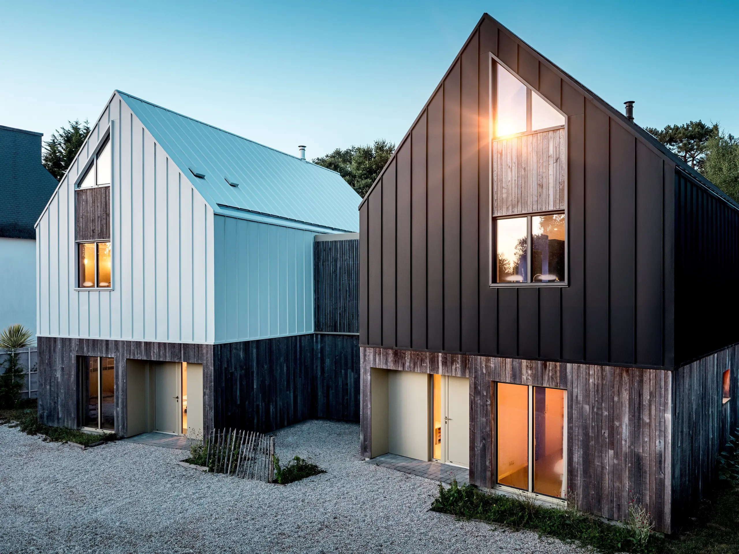 Die beiden Wohnhäuser des Ensembles Le Rhune präsentieren sich mit markanten Satteldächern und kontrastierenden PREFALZ Aluminiumhüllen in P.10 Reinweiß und Schwarzgrau. Die reduzierte Formensprache, inspiriert von traditionellen bretonischen Fischerhäusern, wird durch die präzisen Stehfalzlinien besonders wirkungsvoll betont. Das warme Licht der Innenräume setzt spannende Akzente und verstärkt die architektonische Präsenz der klar gegliederten Baukörper. Die langlebige und wetterbeständige PREFALZ Gebäudehülle verbindet Dach und Fassade nahtlos miteinander, ermöglicht exakte Detailausbildungen und verleiht dem Ensemble eine moderne, hochwertige und zeitlose Ausstrahlung.