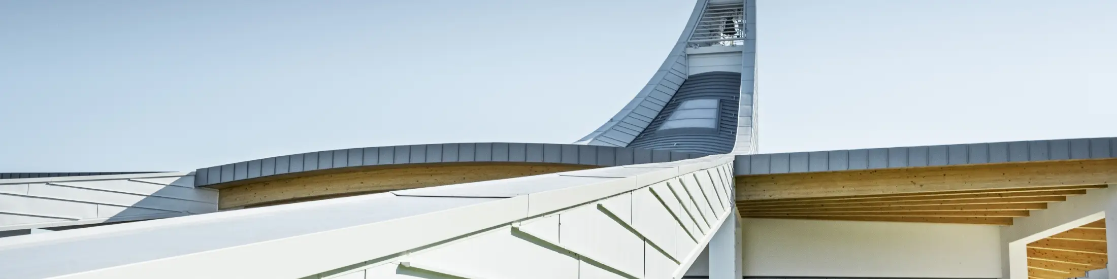 Modernes Kirchengebäude mit geschwungener Dachkonstruktion aus hellen Aluminiumpaneelen. Im Hintergrund erhebt sich ein hoher, elegant gebogener Turm mit einer Statue an der Spitze. Die Architektur kombiniert klare Linien mit offenen Glasflächen und Holzelementen unter dem Dach. Der Himmel ist wolkenlos und betont die helle, futuristische Gestaltung des Bauwerks