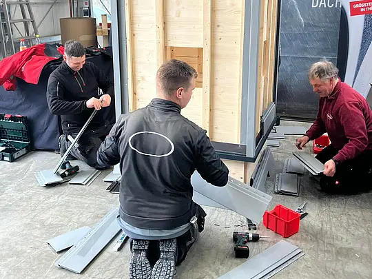 Vor-Ort-Training mit PREFA Academy Cube: Drei Handwerker montieren Sidings an eine Trainingskonstruktion in einer Werkhalle. Die Fachkräfte arbeiten kniend an einer Holzunterkonstruktion und verarbeiten vorgefertigte Aluminium-Sidings. Am Boden liegen Werkzeuge wie Akkuschrauber und Schrauben, während im Hintergrund ein PREFA-Branding und die Industriehalle sichtbar sind.