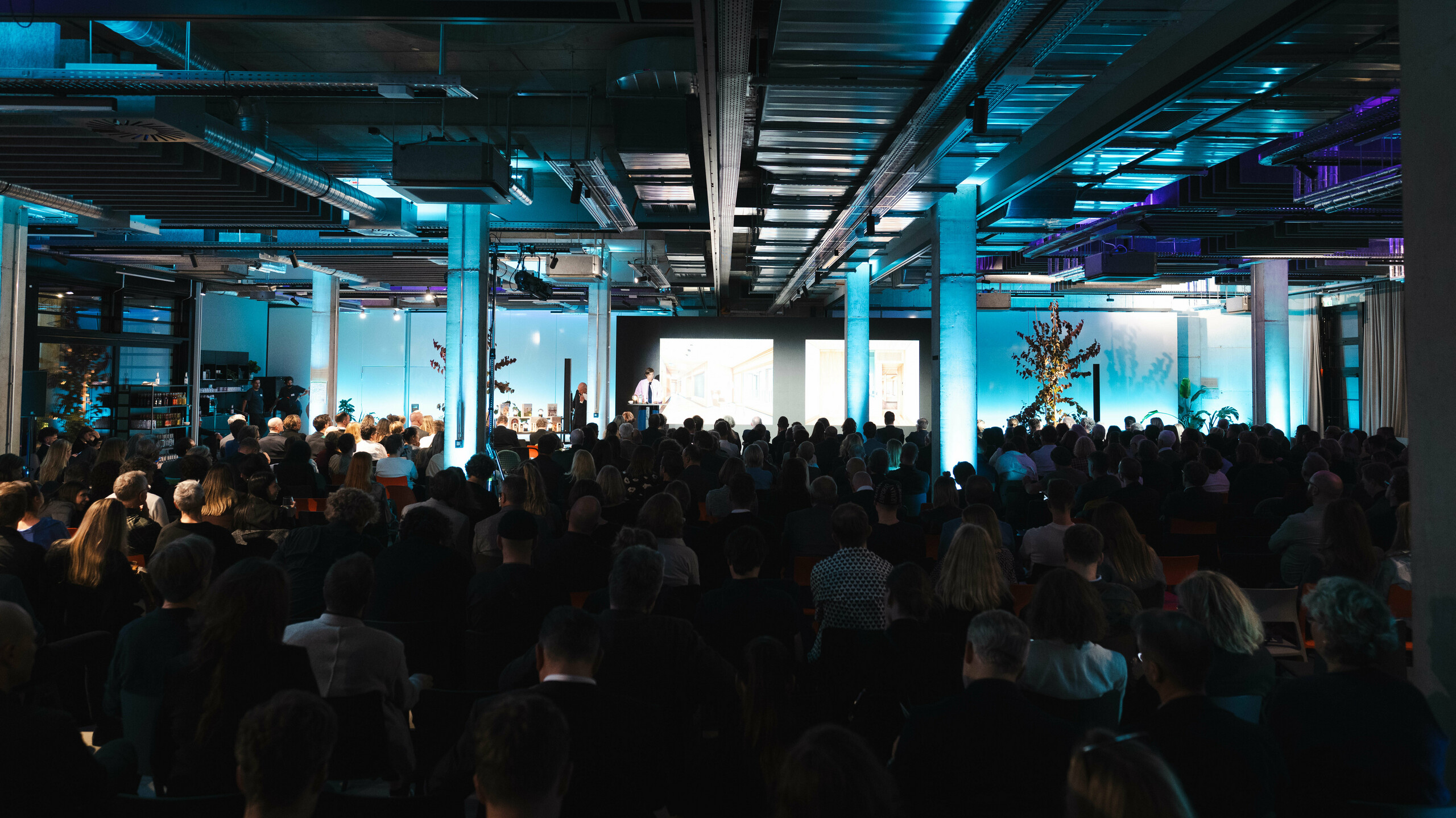 Atmosphärische Event-Szene bei der Preisverleihung ‚Wohnbauten des Jahres‘ 2025: Blick von hinten auf ein voll besetztes Auditorium in einer modernen Industriehalle mit auffälliger blauer Beleuchtung. Im Vordergrund die Bühne mit großer Leinwand, auf der Präsentationen gezeigt werden. Die Architektur des Raumes mit sichtbaren Stahlträgern und Betonpfeilern unterstreicht den innovativen Charakter des Events, das nachhaltige Lösungen wie das Solarmodul PREFALZ feiert.